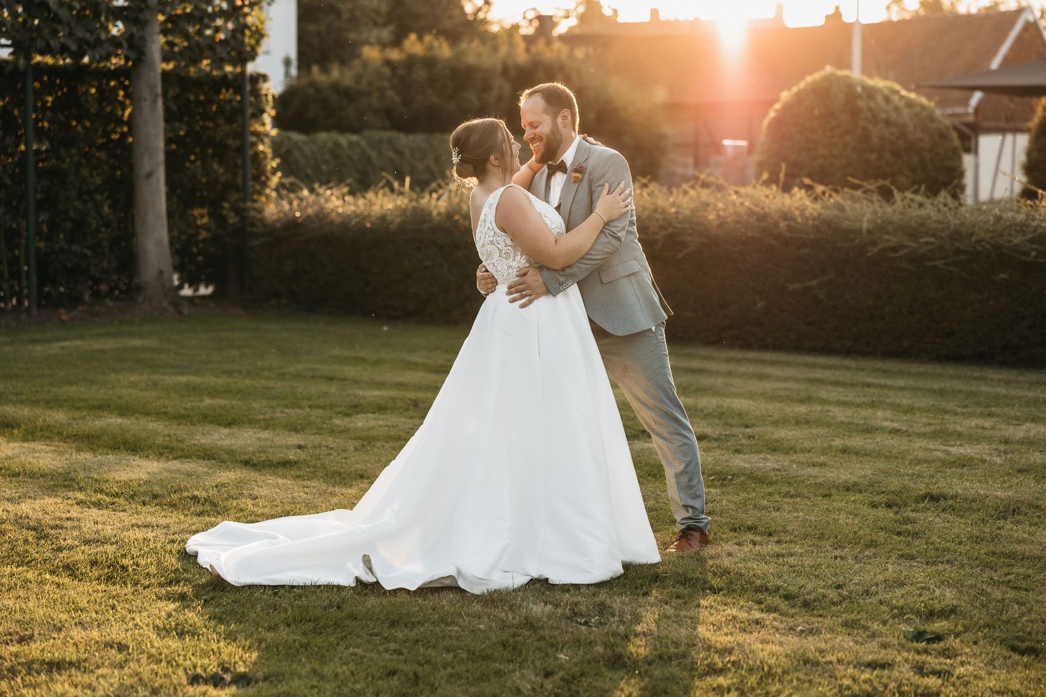 Newlywed couple embracing and kissing in garden with warm golden hour sunset light.