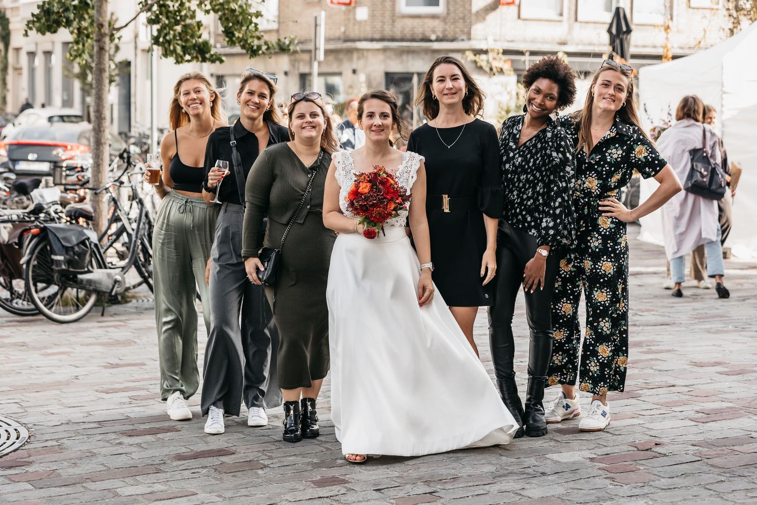 Bride posing with six bridesmaids on a cobblestone street holding a red bouquet.