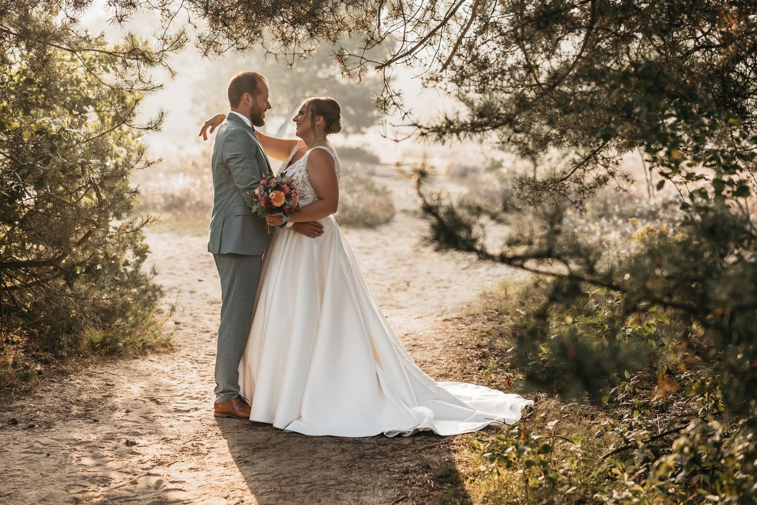 Bride and groom share a romantic moment on a sandy path surrounded by lush trees in golden sunlight.