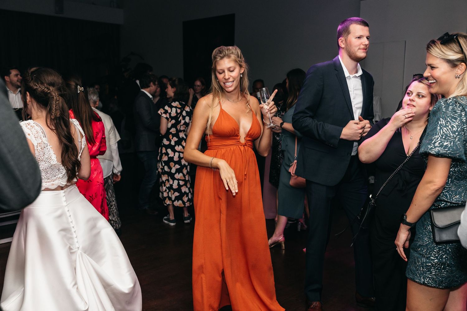 Woman in orange dress dances near bride at elegant wedding reception celebration.