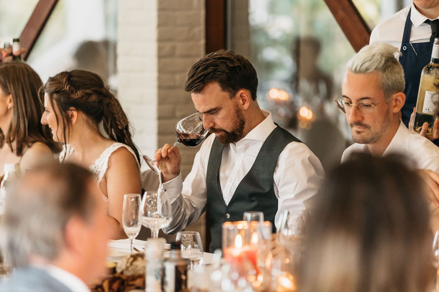 Man in vest sipping red wine at an elegant wedding reception dinner table.