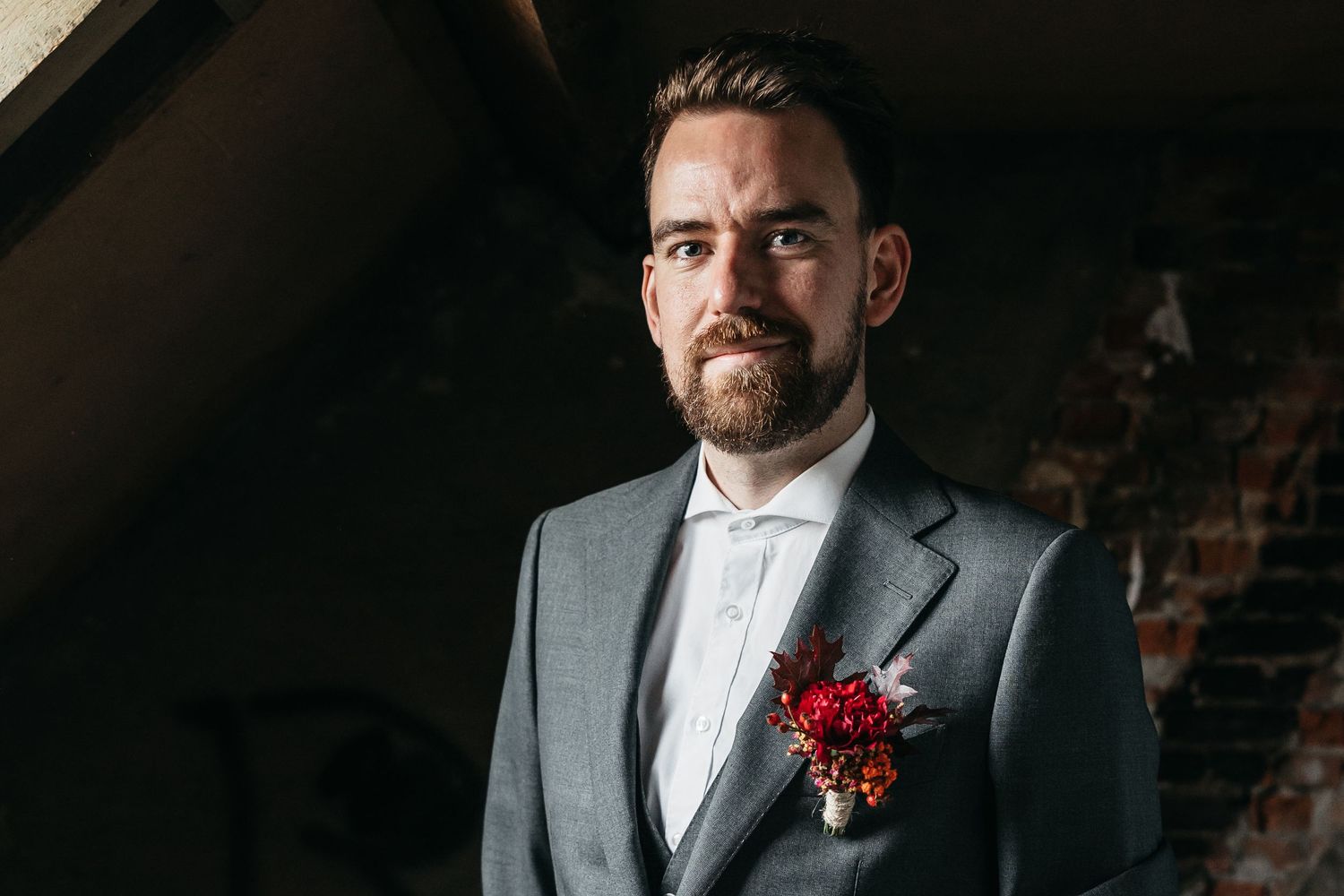 Serious bearded groom in gray three-piece suit with red floral boutonniere in rustic venue.