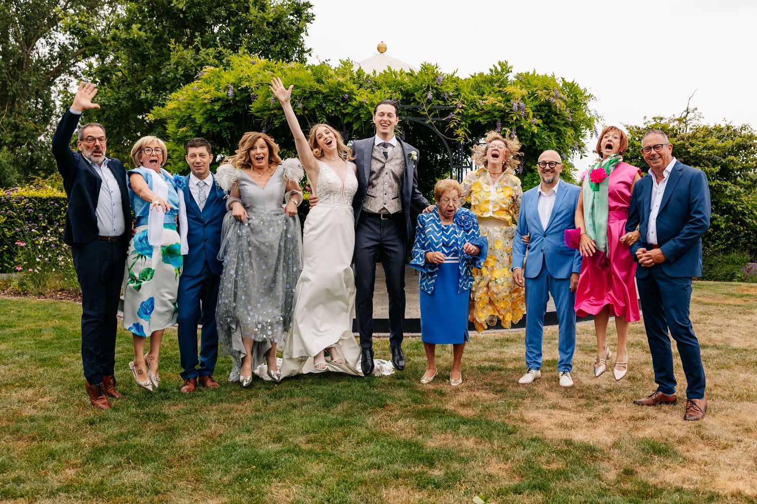 A joyful wedding group including bride, groom, and family celebrates outdoors with arms raised in cheer.