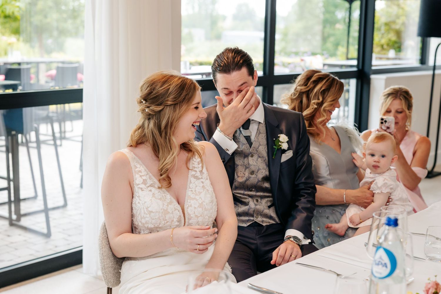 Emotional groom covers his mouth while bride smiles during wedding reception speeches.