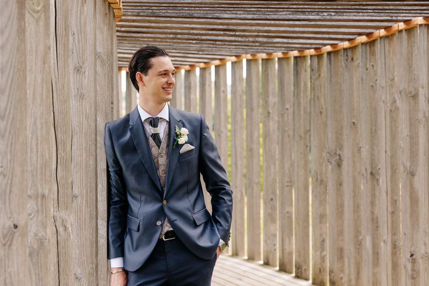 Handsome groom in navy suit with boutonniere smiling while leaning against wooden structure.