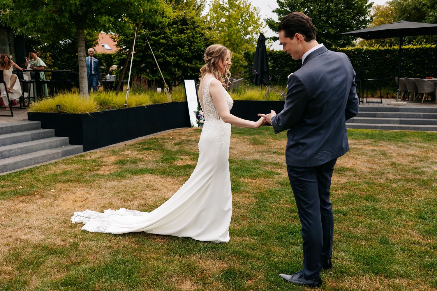 Bride in elegant white gown reads vows to groom during outdoor first look ceremony on lush green lawn.