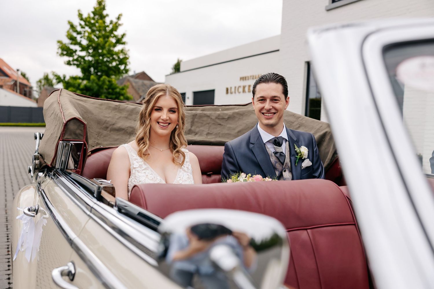 Bride and groom posing joyfully in a vintage cream convertible car outside Blickenberg wedding venue.