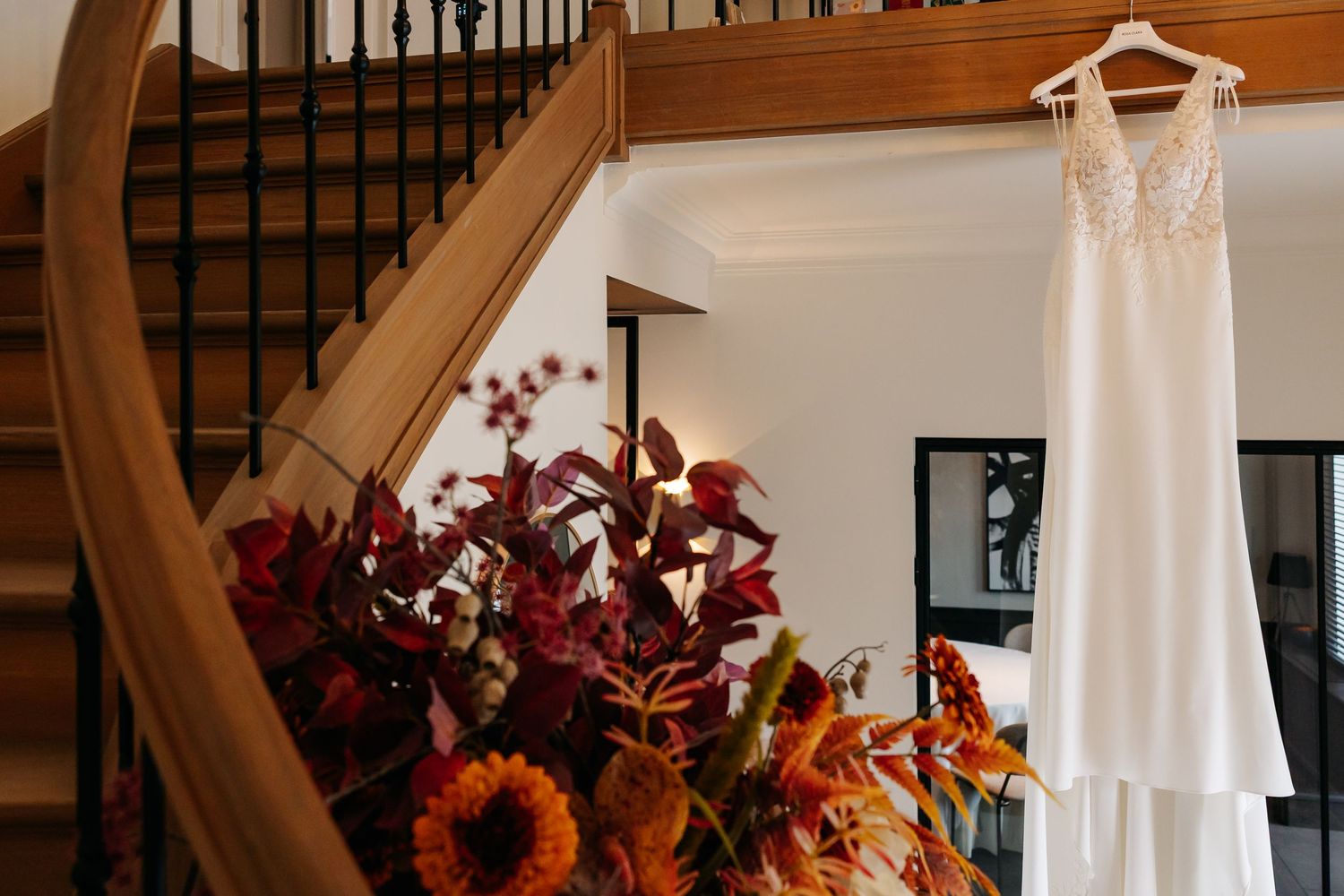 Elegant white lace wedding dress hanging on staircase beside vibrant autumn floral arrangement.