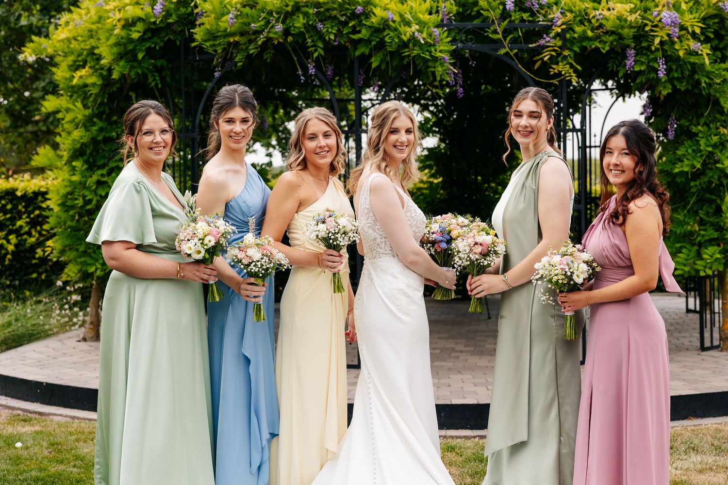 Bride in sleeveless lace gown poses smiling with five bridesmaids in pastel green, blue, and pink dresses.
