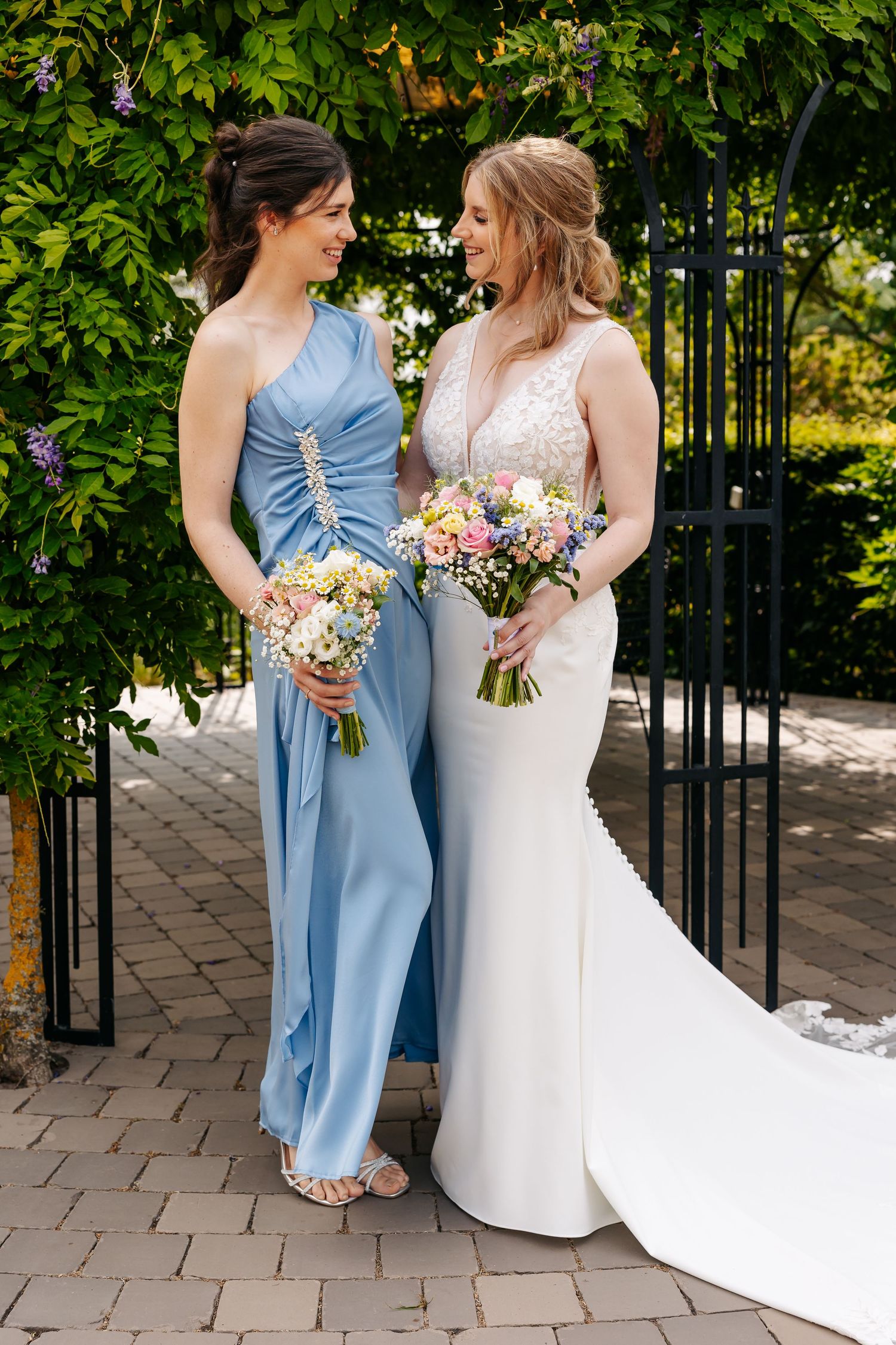 Bride and bridesmaid in blue satin dress smile at each other while holding bouquets under a leafy garden arch.