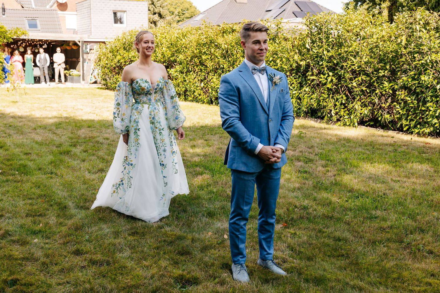 Bride in floral wedding gown and groom in blue suit stand on green lawn during outdoor wedding ceremony.