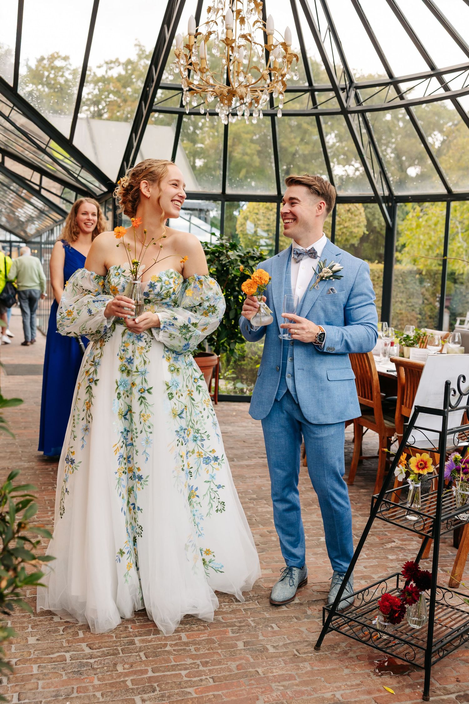 Bride in floral wedding gown and groom in blue suit walking through glass greenhouse wedding venue.