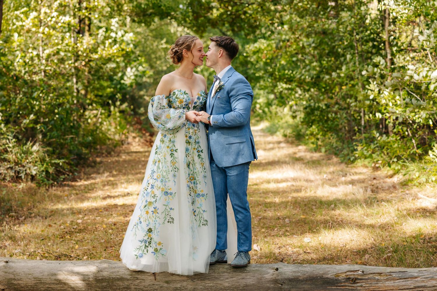 A bride and groom hold hands and gaze at each other on a sunlit forest path during their wedding.