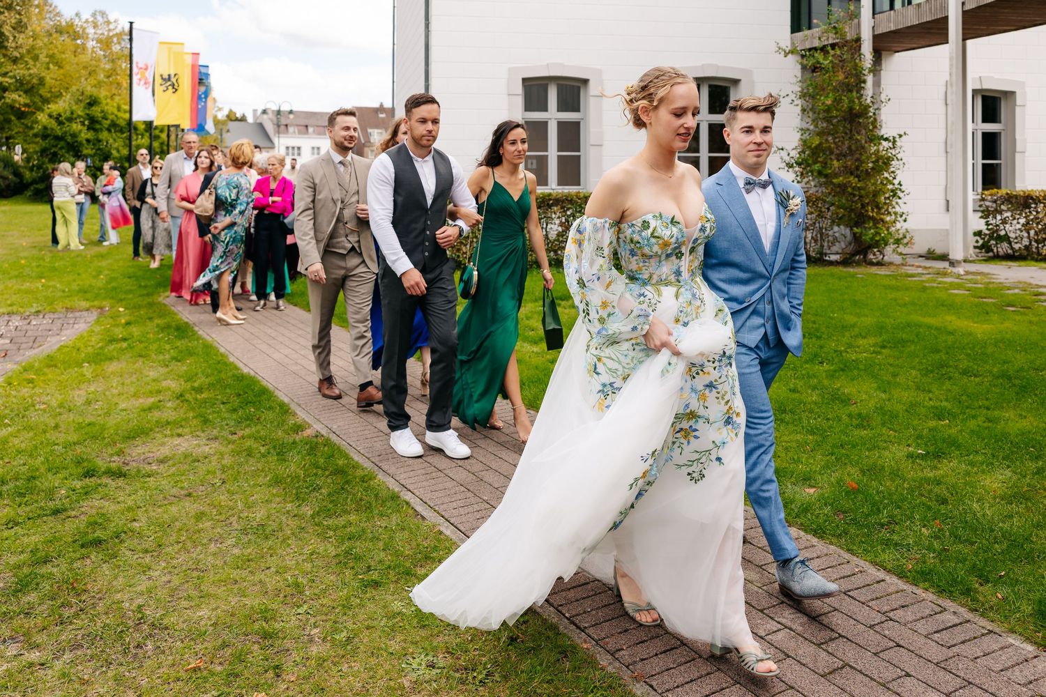 Wedding party procession led by bride in floral gown and groom in blue suit.