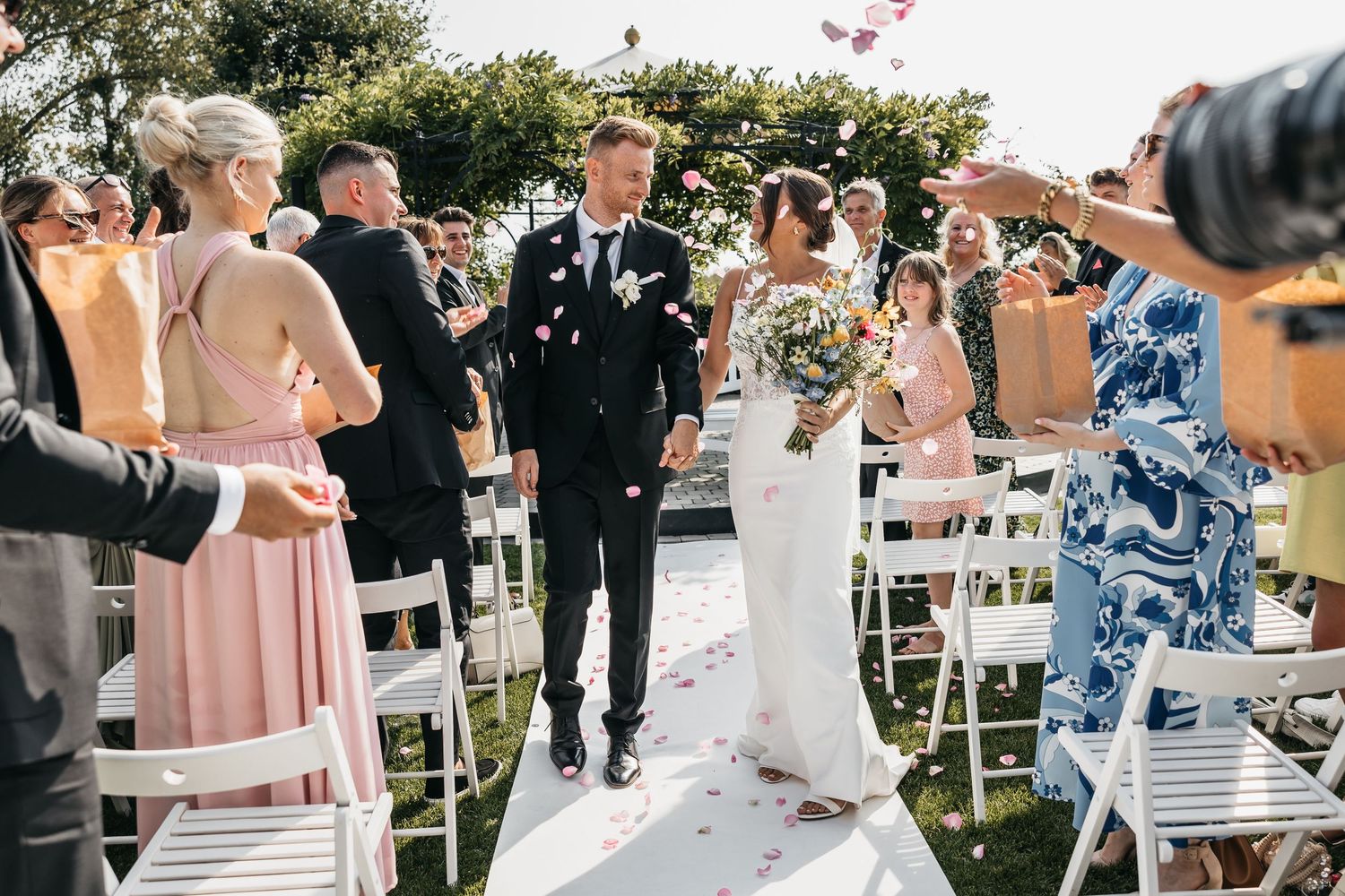 Newlywed couple walks down the aisle surrounded by guests throwing confetti at outdoor ceremony.