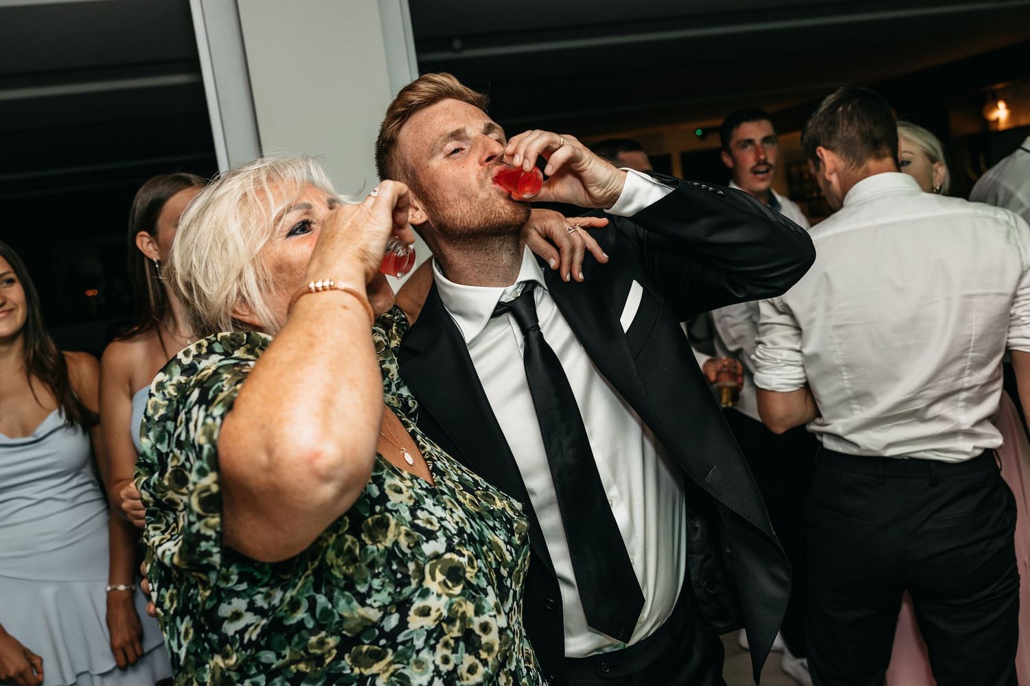 Groom and older female guest take celebratory shots together during fun wedding reception party.