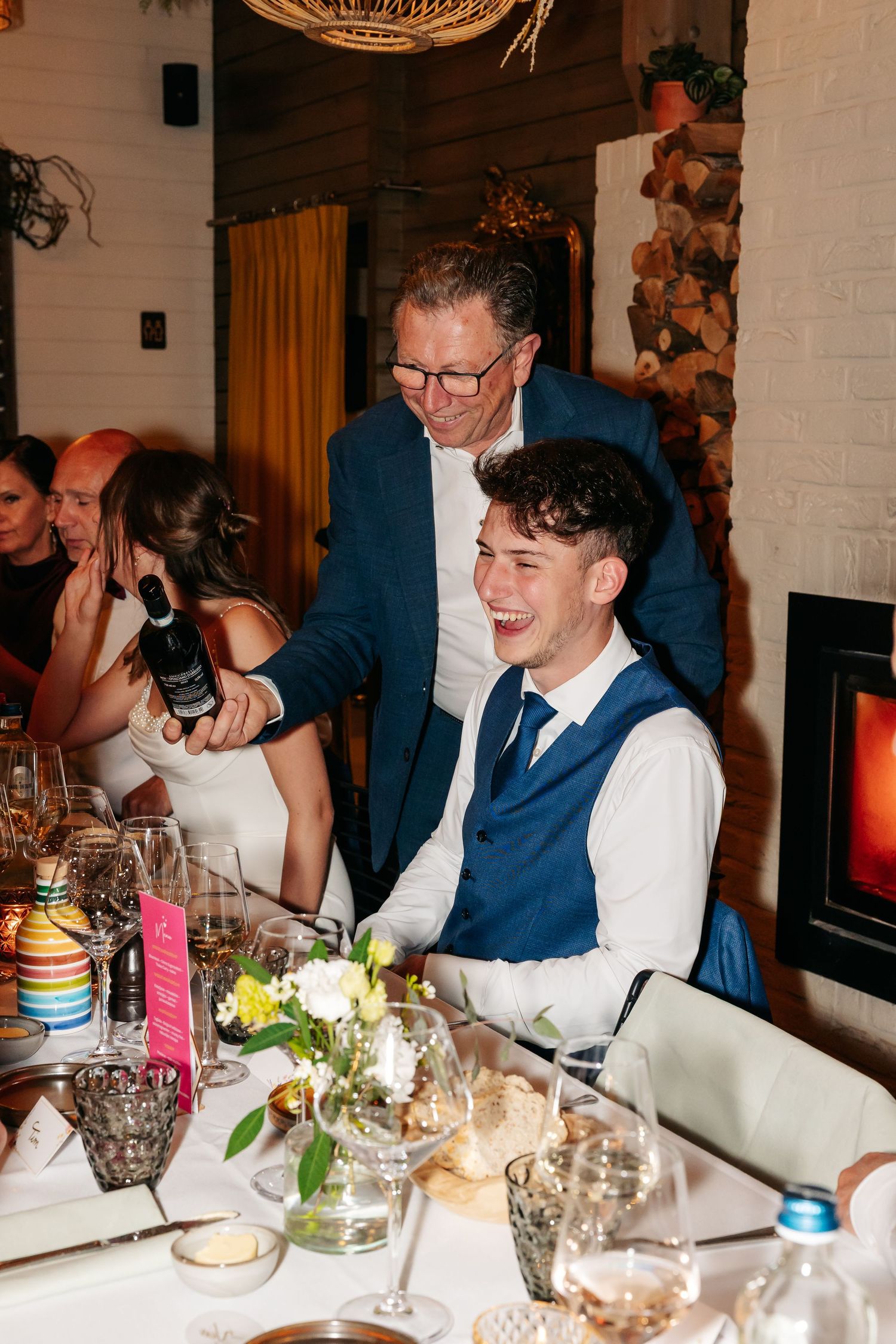 Older man pouring wine for laughing young guest at elegant wedding reception dinner table.
