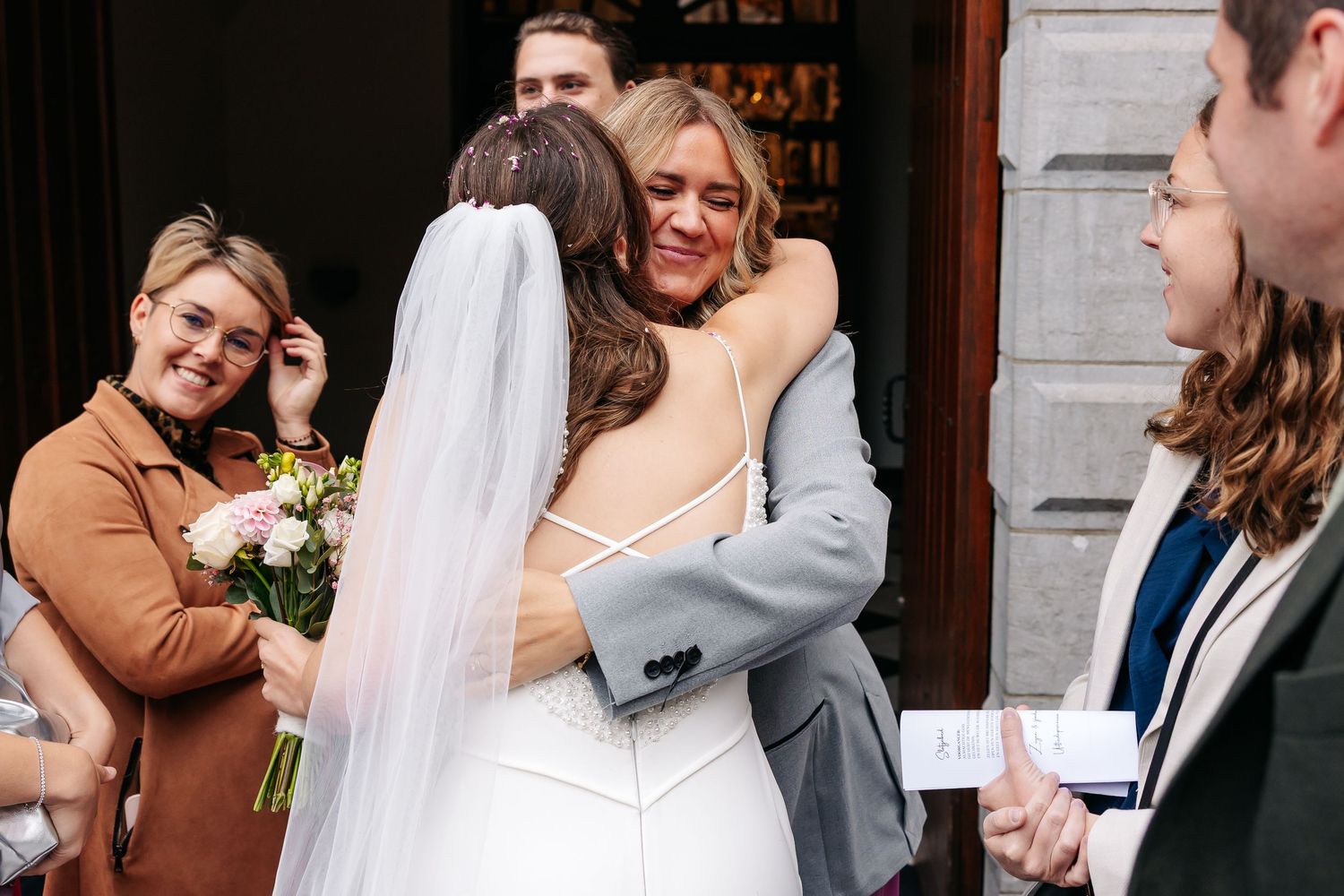 Bride in white gown and veil shares an emotional hug with a smiling female guest outside the church.