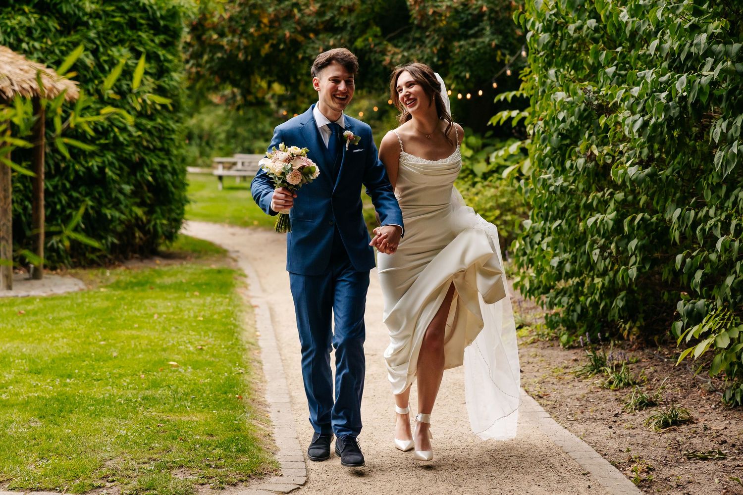 Smiling bride and groom walking hand in hand along a sunlit garden path, groom holding a floral bouquet.