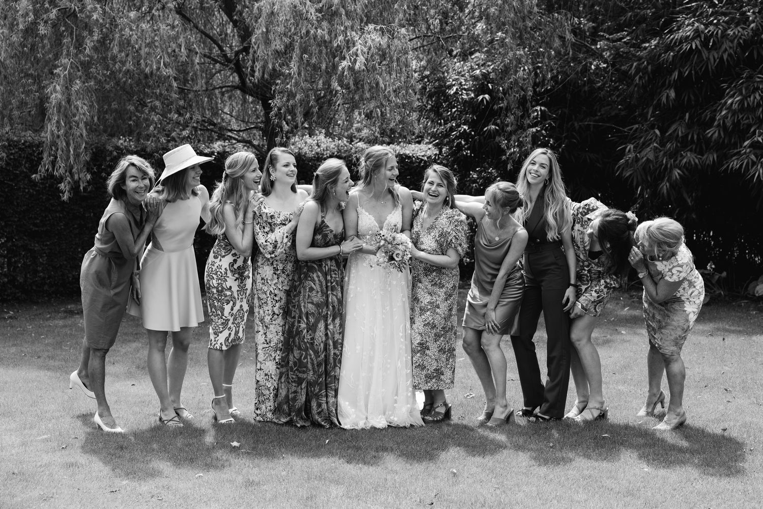 Black and white photo of bride with bridesmaids laughing outdoors under a willow tree.