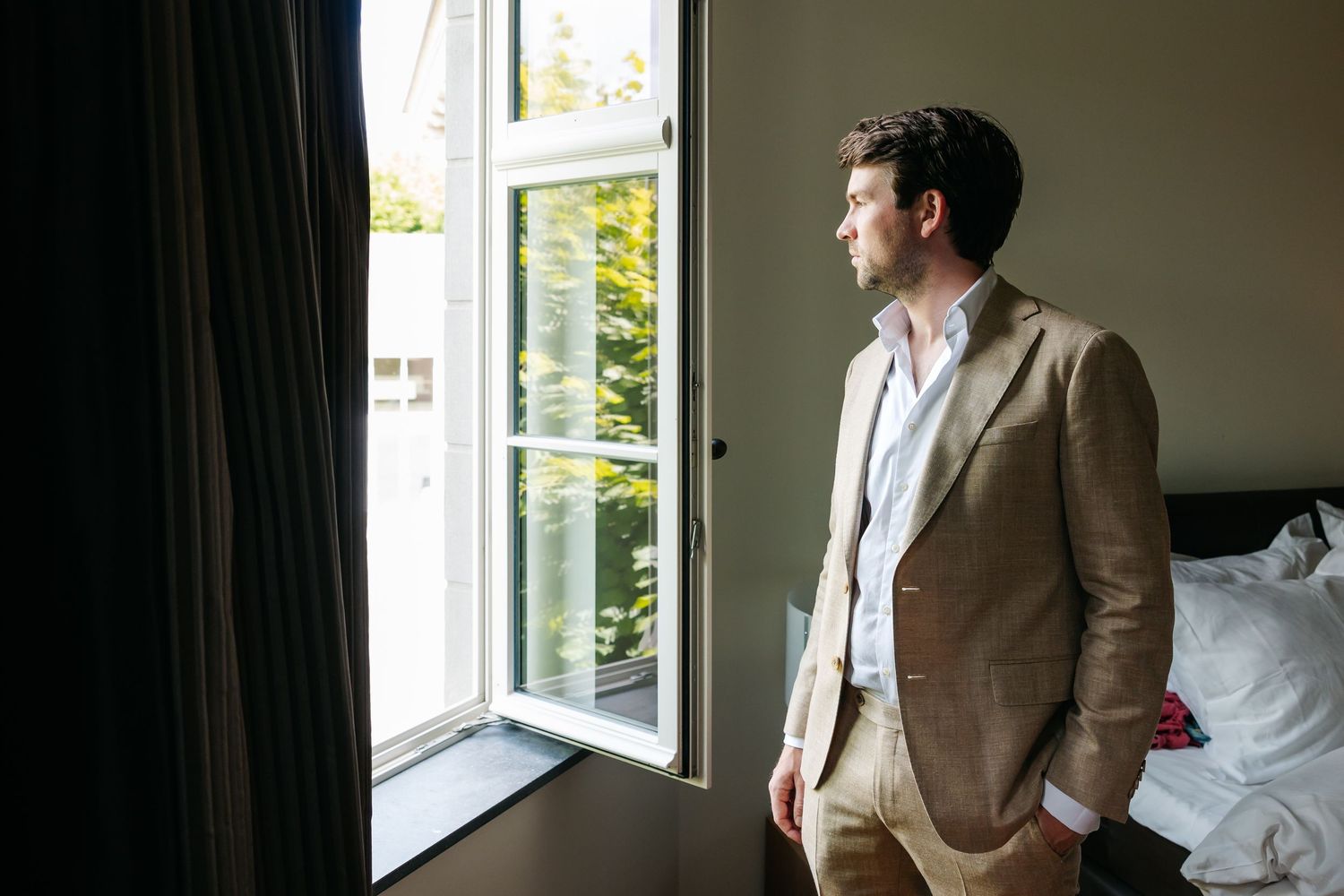 Groom in tan suit gazing out open window in hotel room before wedding ceremony.