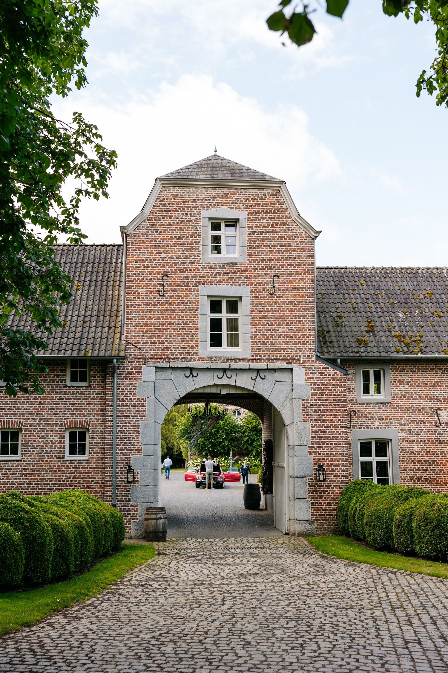 Historic brick gatehouse with arched entrance, cobblestone driveway, and manicured hedges at a Belgian estate venue.