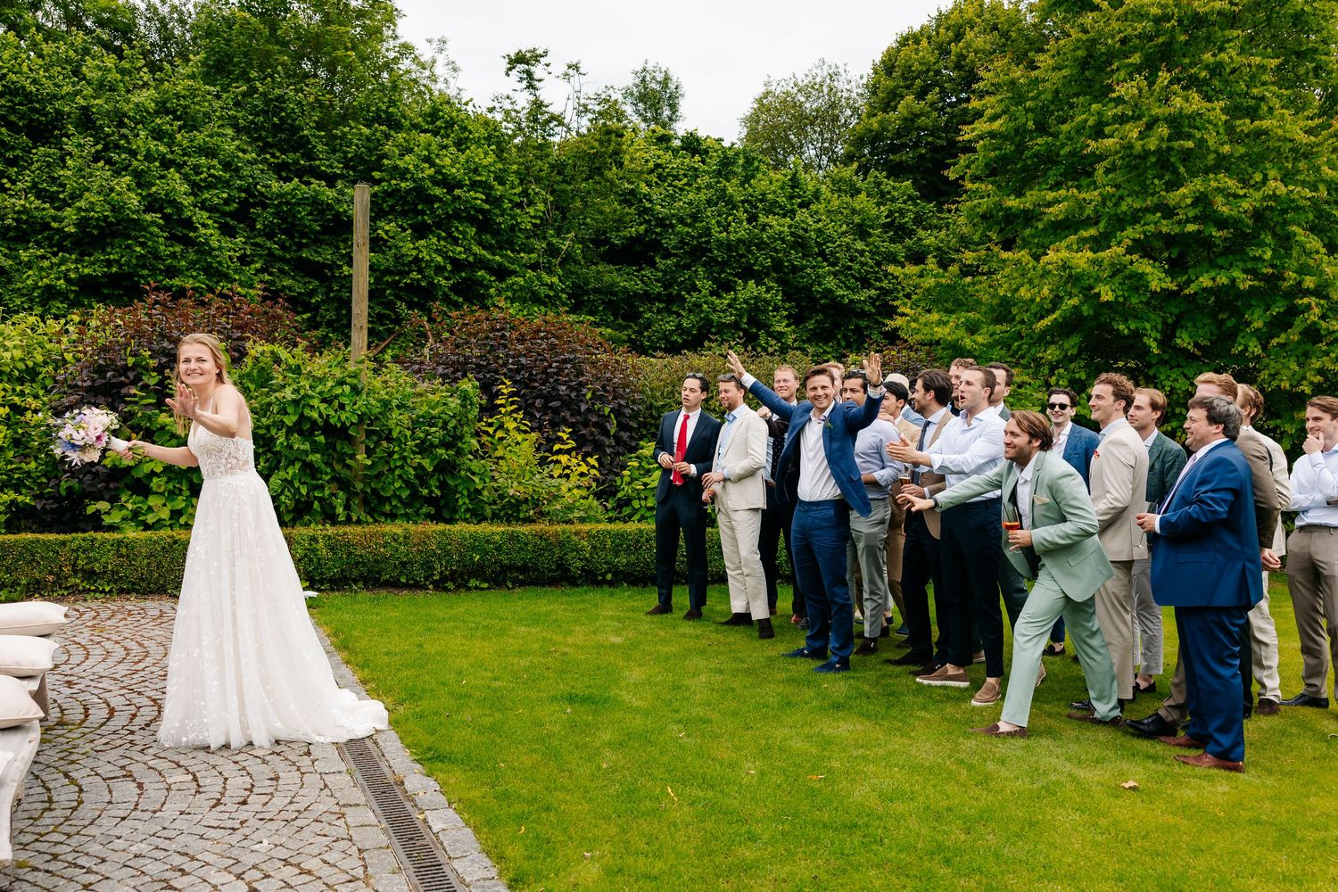 Bride in white gown prepares to toss bouquet to group of male wedding guests in garden.
