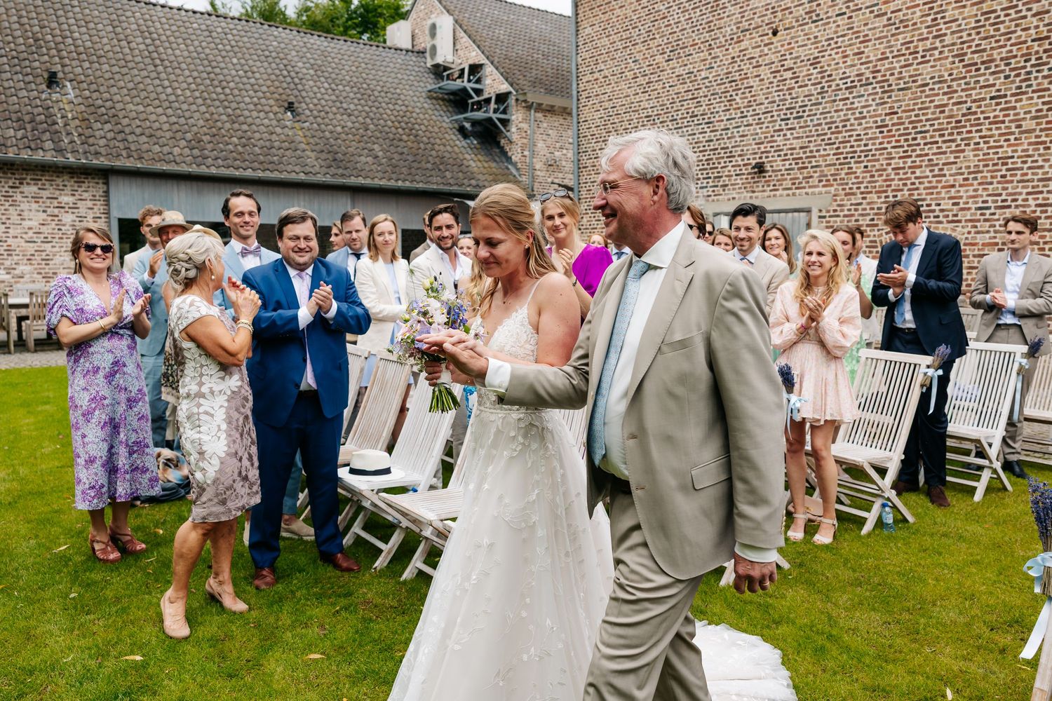 Bride in white gown walking down outdoor aisle with older man while guests applaud at sunny wedding.