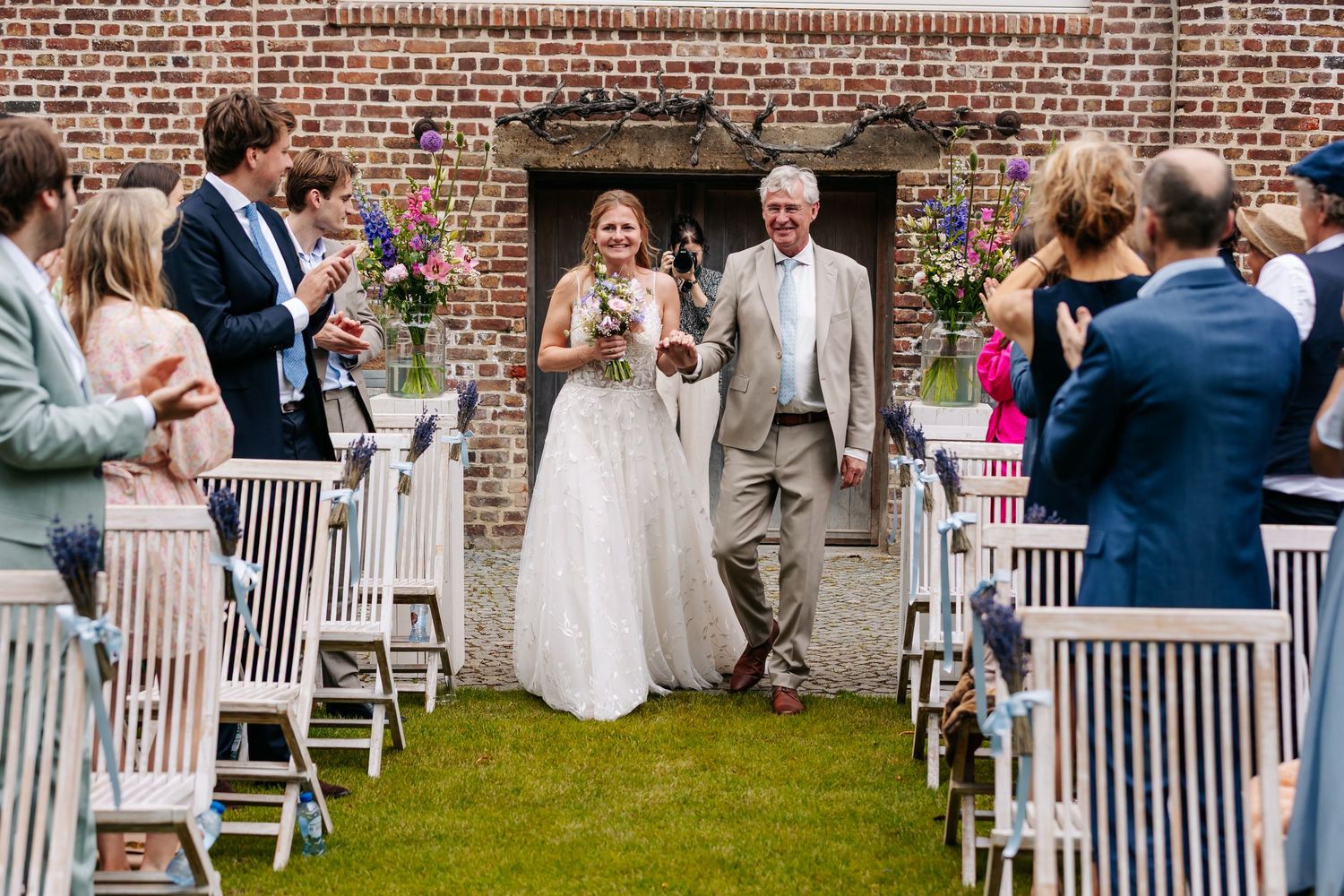Bride in white gown walking down aisle with father at outdoor garden wedding ceremony.