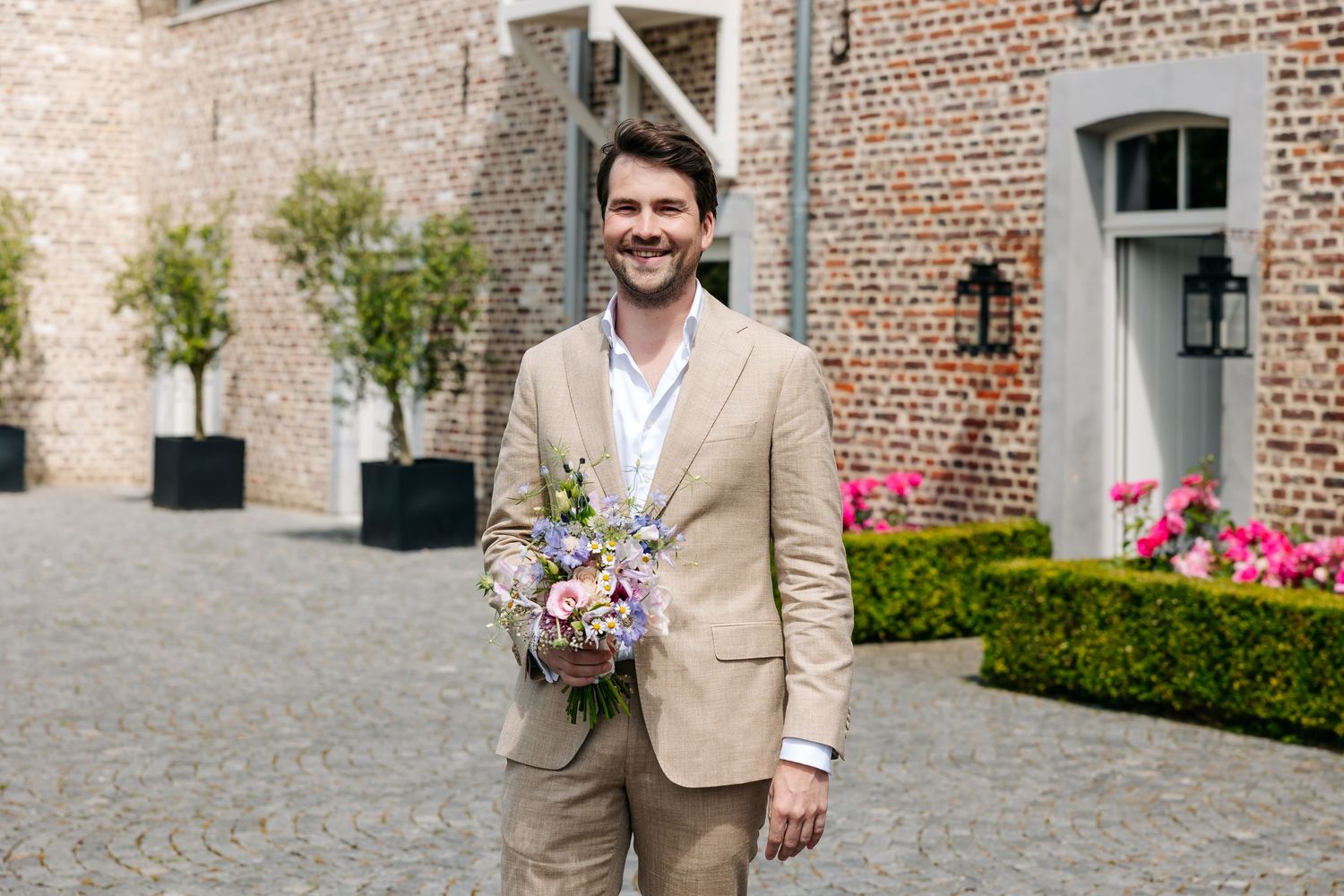 Smiling man in a beige suit holding a colorful bouquet on a cobblestone courtyard outside a brick building.
