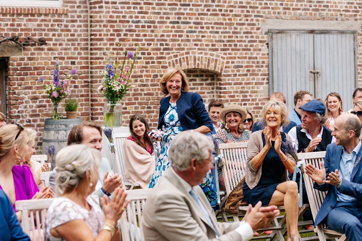 Wedding guests applauding speaker at sunny outdoor ceremony beside rustic brick wall with florals.