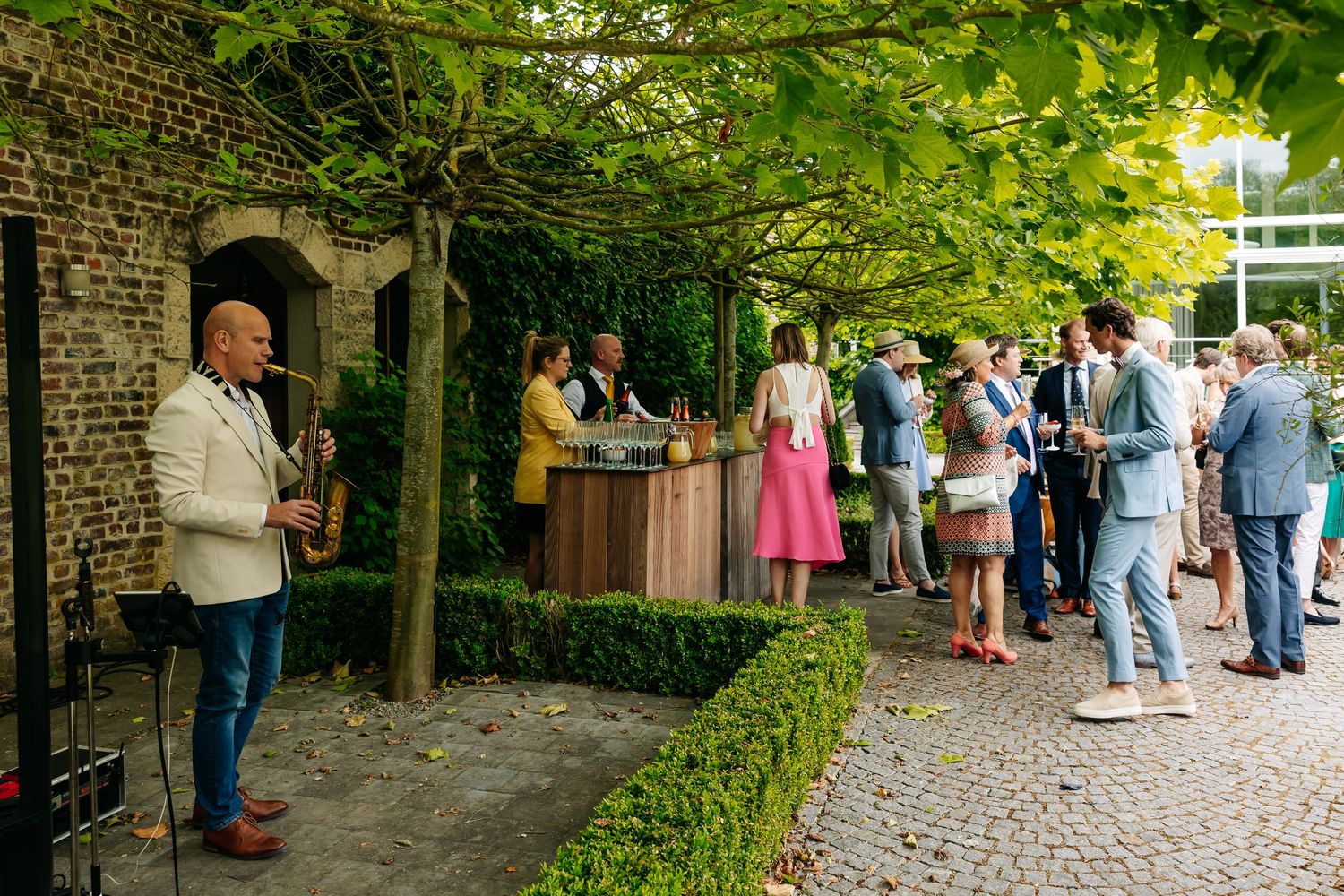 Saxophonist performing at outdoor wedding reception while guests mingle near bar under green vine canopy.