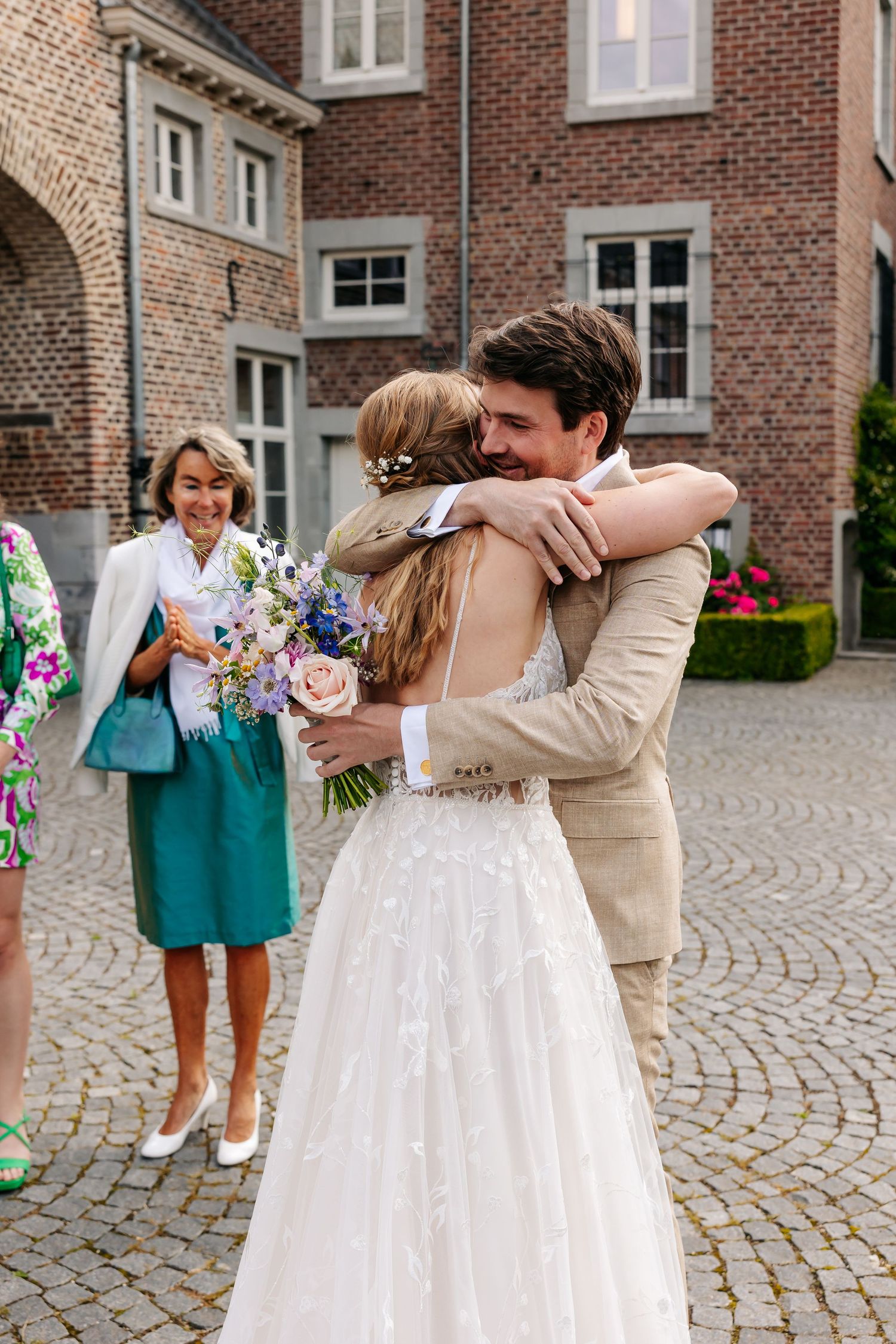 Groom in beige suit embracing bride in lace wedding dress holding colorful bouquet in courtyard.
