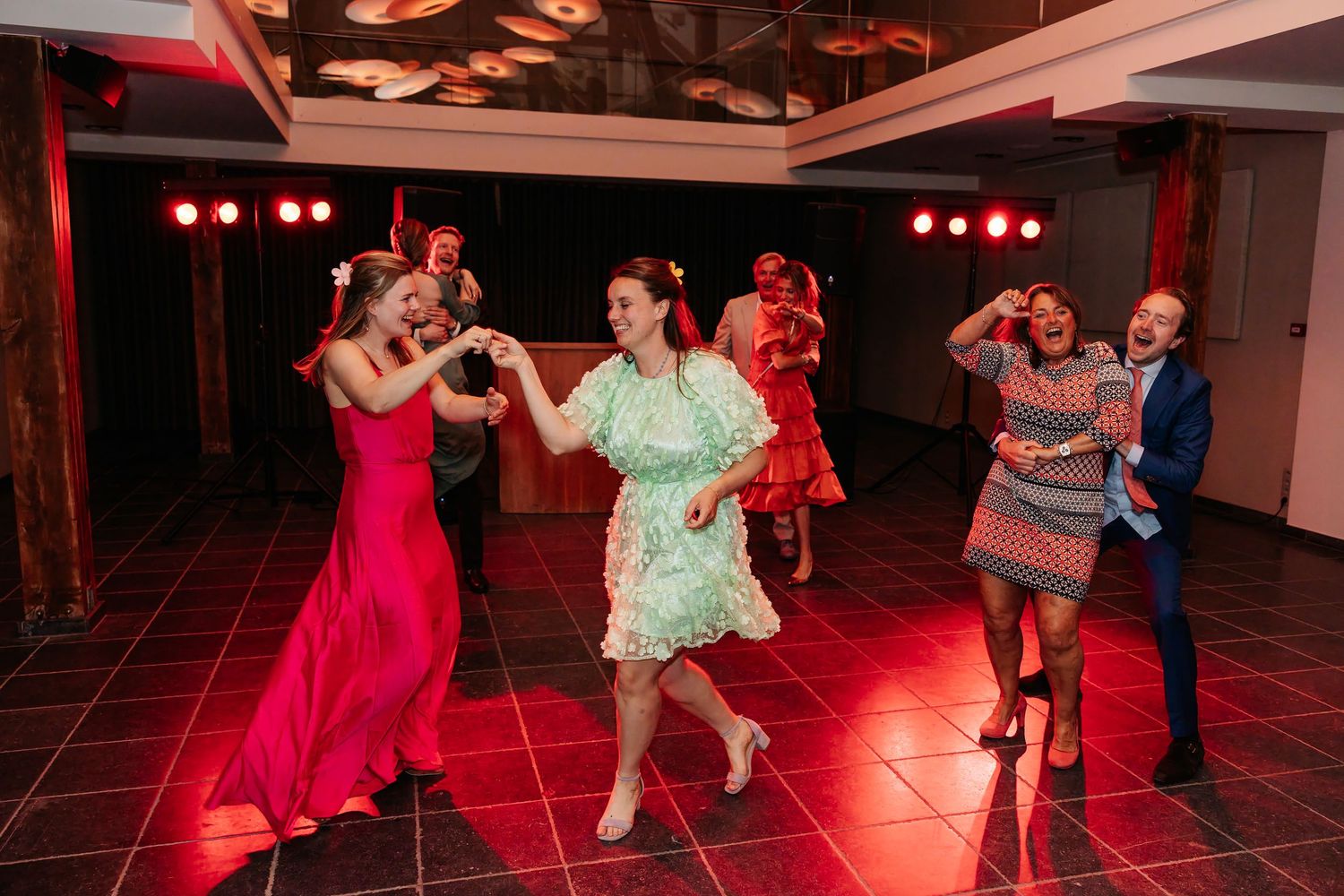Energetic wedding guests dancing under red lights, women in green, pink, and floral dresses on dance floor.