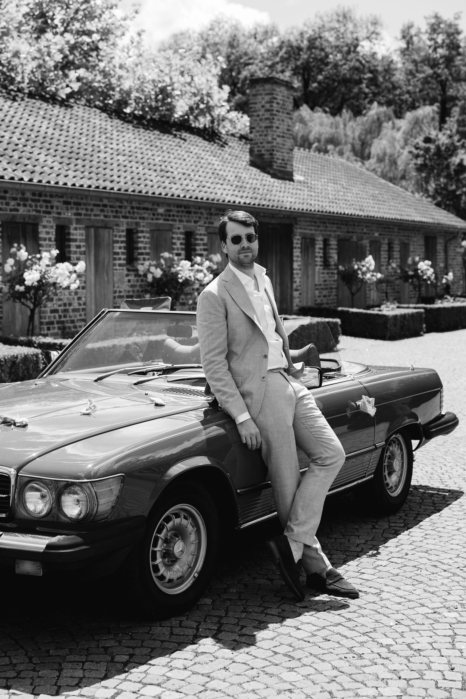 Groom in beige suit leaning against vintage red convertible car in black and white photo outside brick venue.