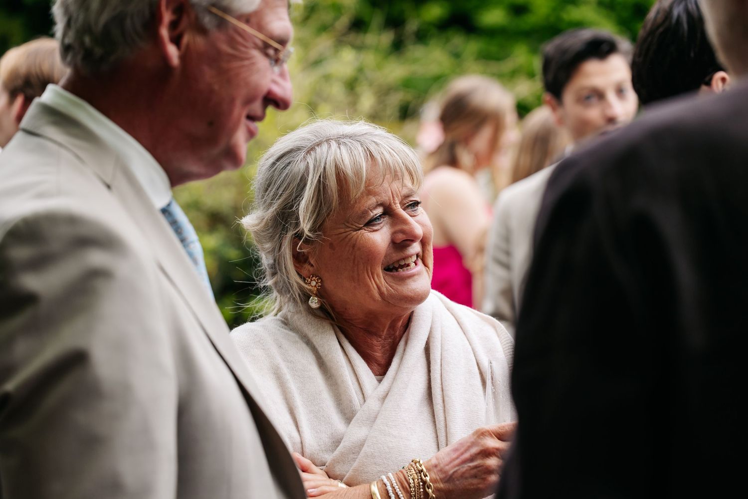 Elegant older woman smiling warmly at wedding reception guest in sunlit garden setting.