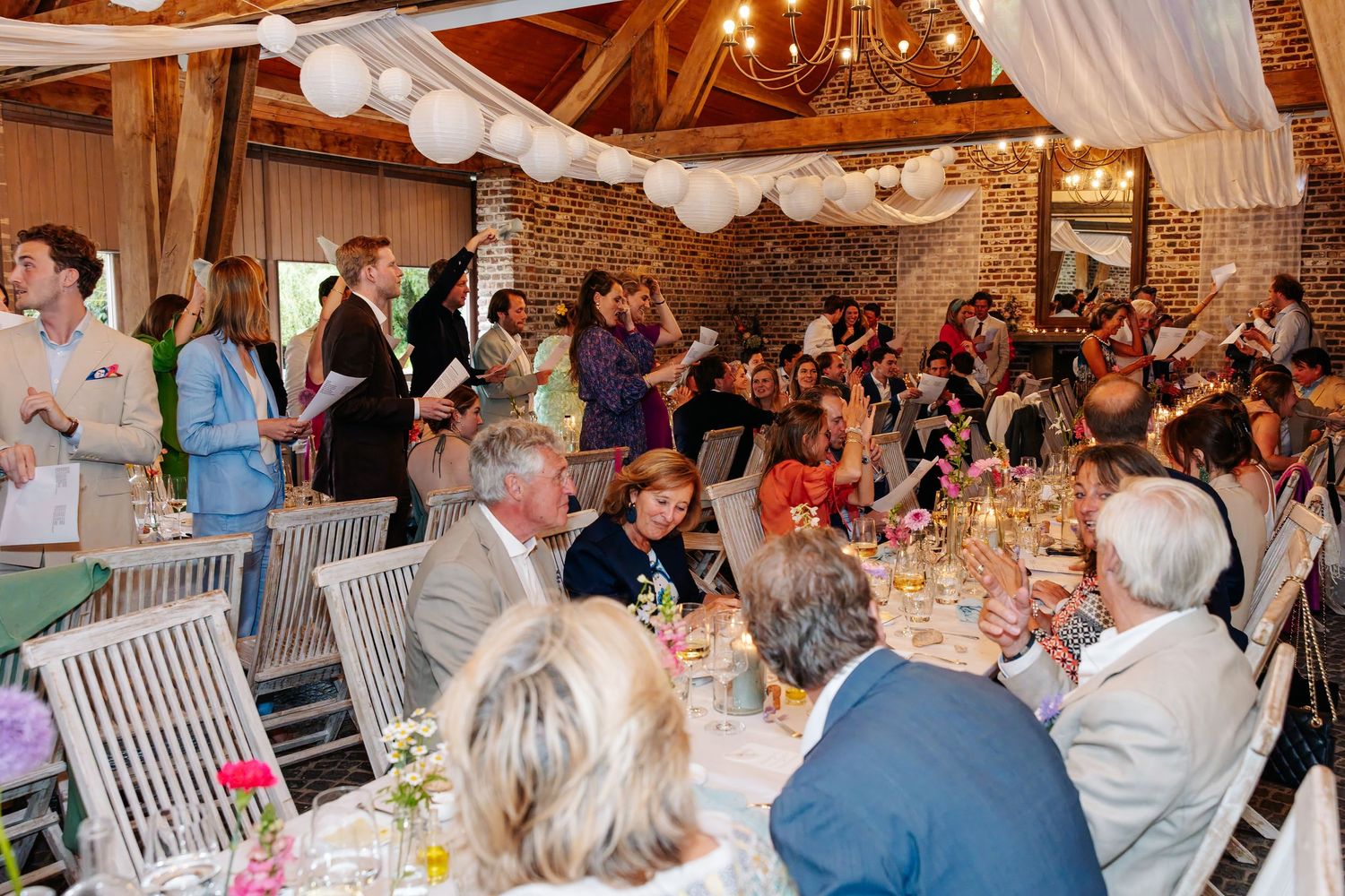 Guests celebrate and raise hands at elegant wedding reception inside warmly lit rustic barn venue.
