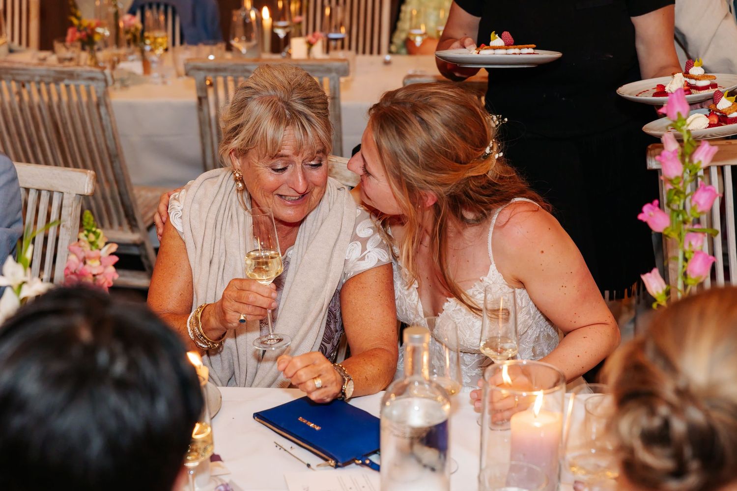 Two women share a joyful moment over champagne at a candlelit wedding reception table.