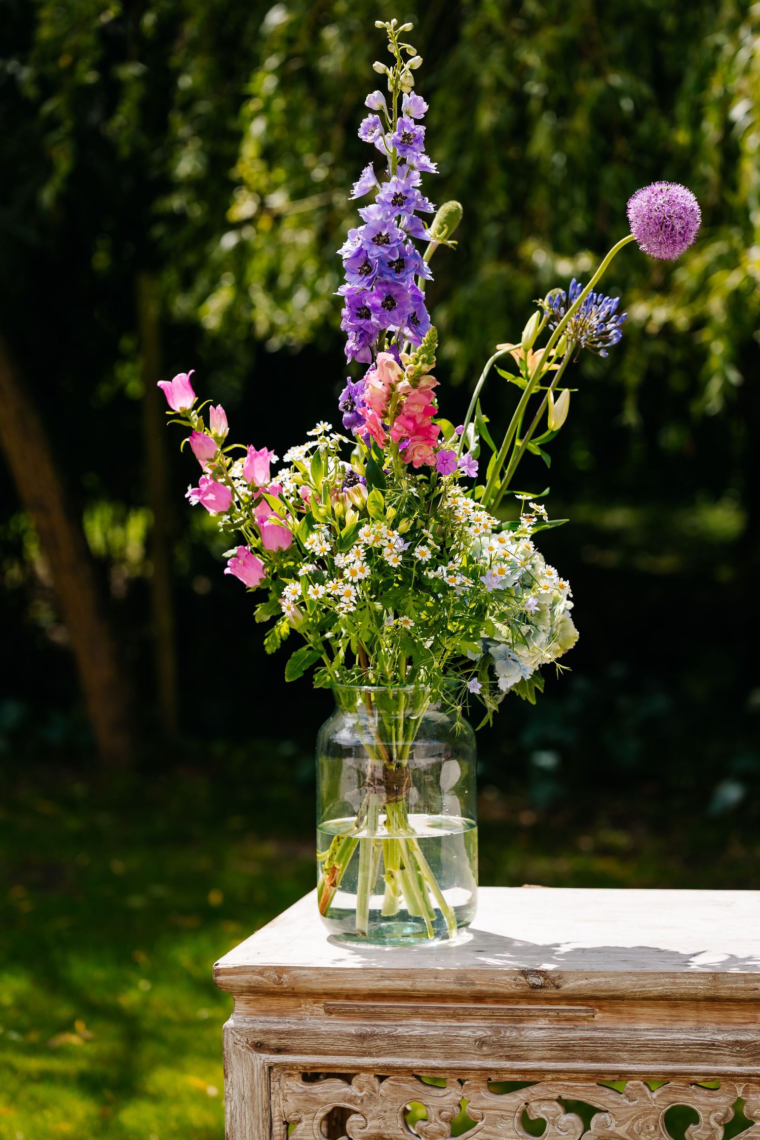 Colorful wildflower bouquet with purple delphiniums and pink blooms in glass vase outdoors.