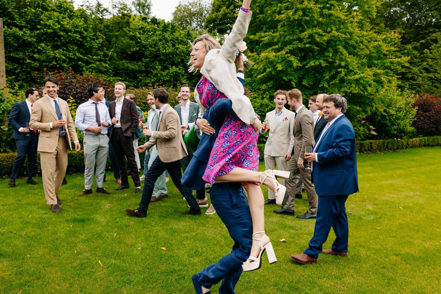 Wedding guest being lifted joyfully by another guest while celebrating on lush green lawn.