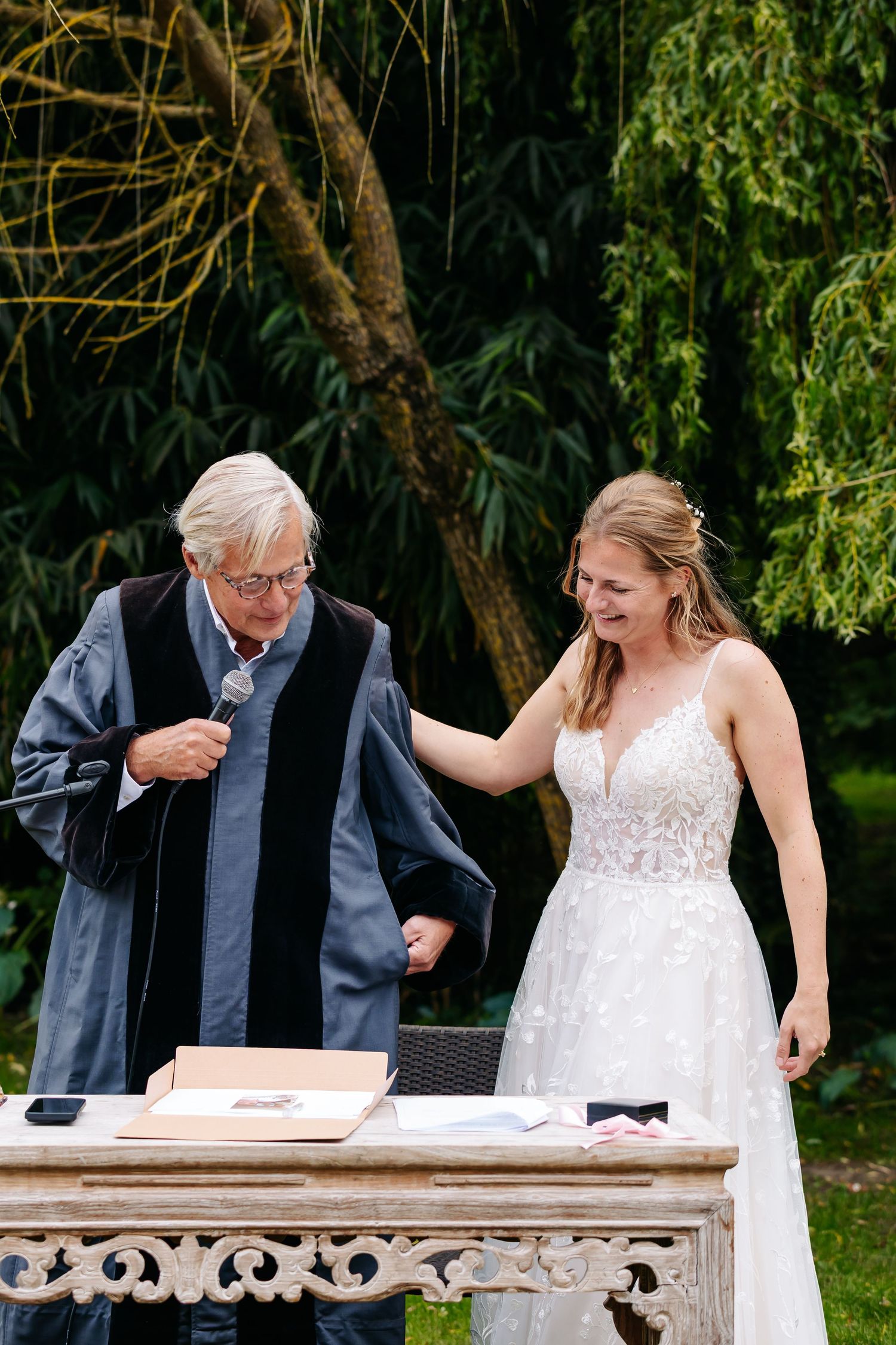 Bride in lace wedding gown laughs with elderly officiant during outdoor wedding ceremony at decorated altar.
