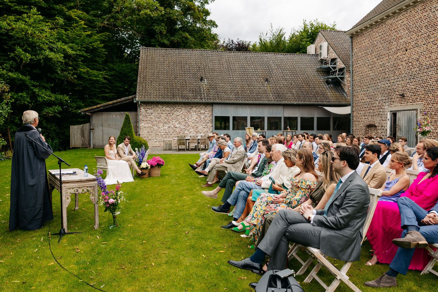 Outdoor wedding ceremony with guests seated on white chairs on a green lawn.