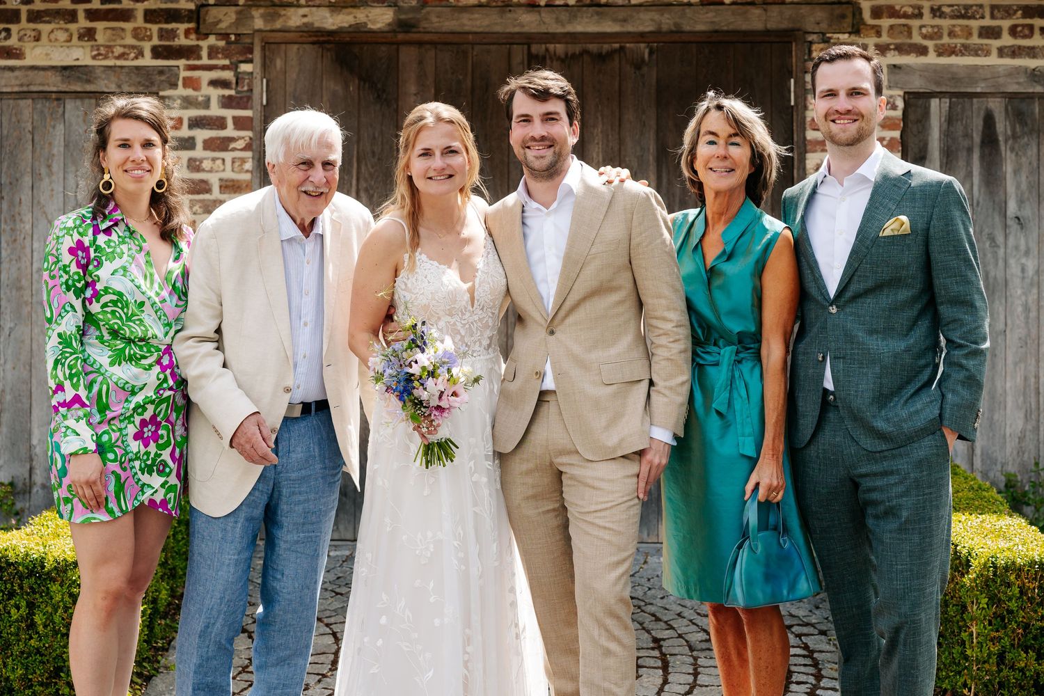 Happy wedding party group photo with bride in lace gown and groom in beige suit outdoors.