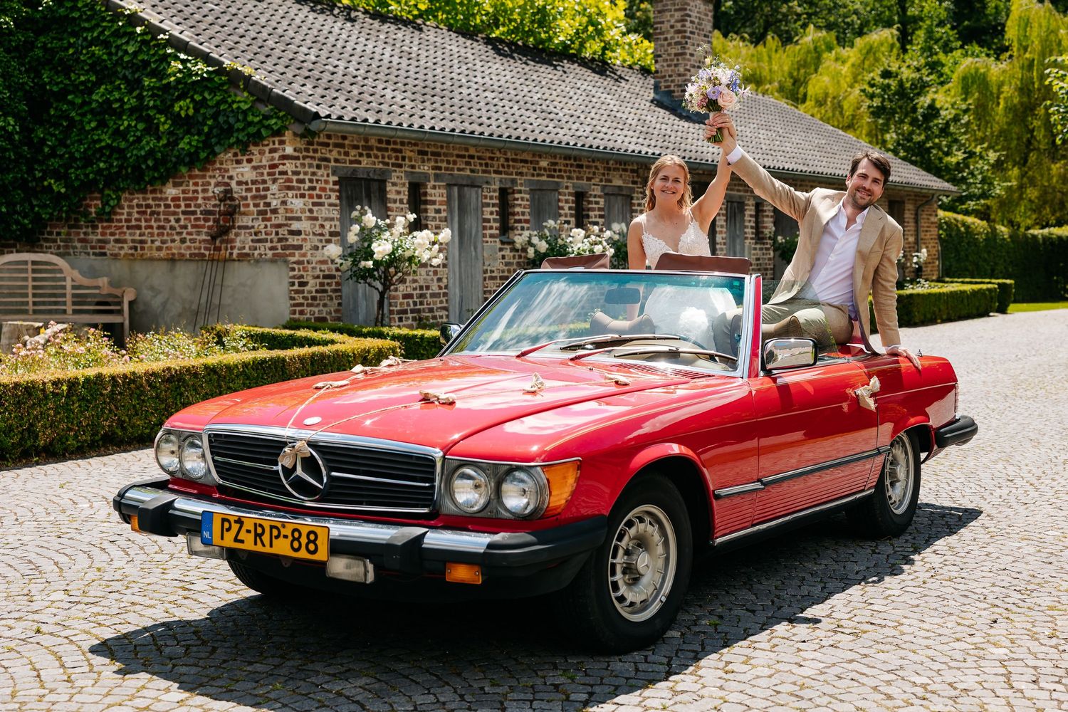 Bride and groom celebrate in red vintage Mercedes convertible on cobblestone driveway.