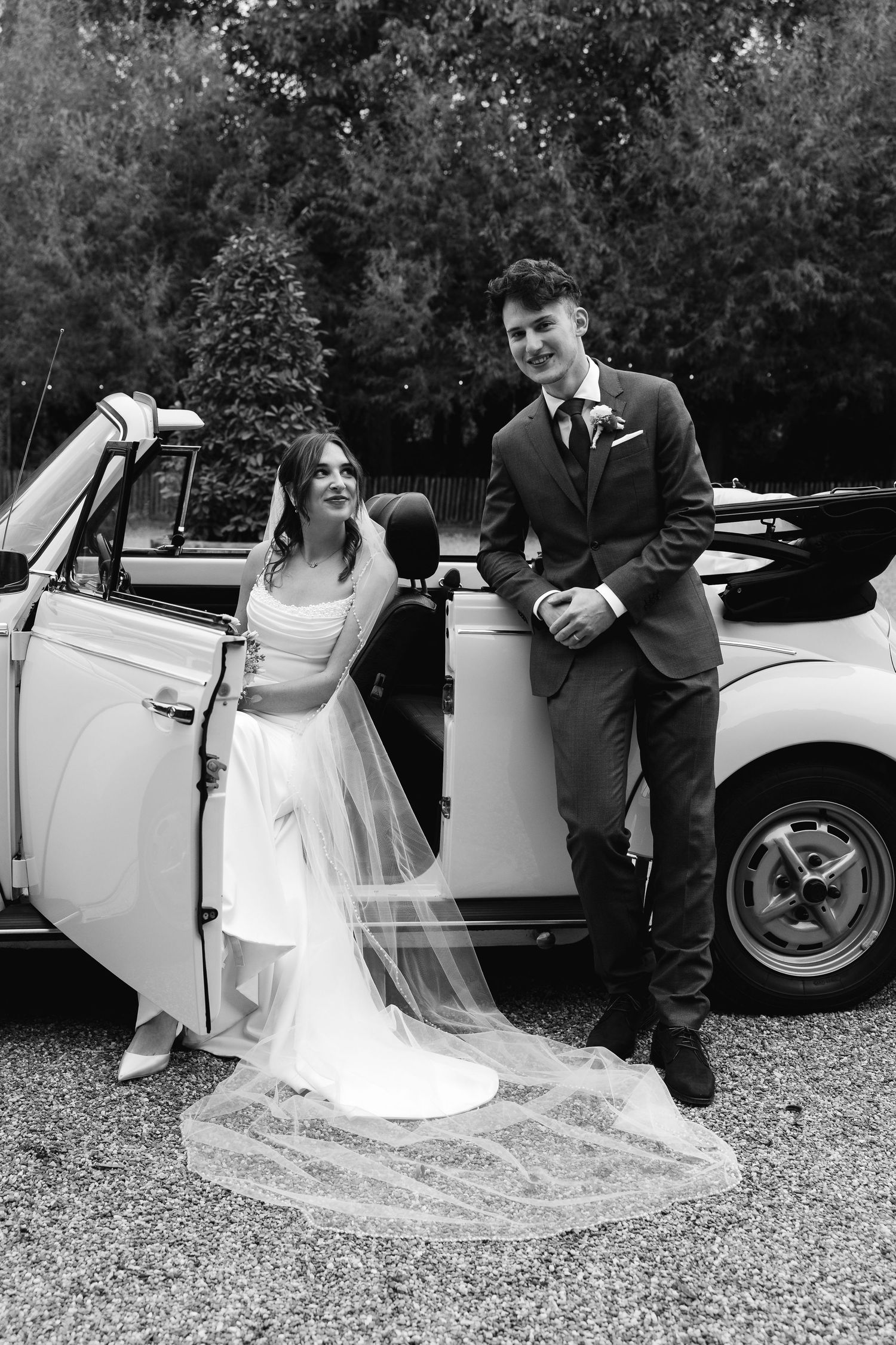 Black and white portrait of smiling bride and groom posing beside a classic white convertible wedding car.