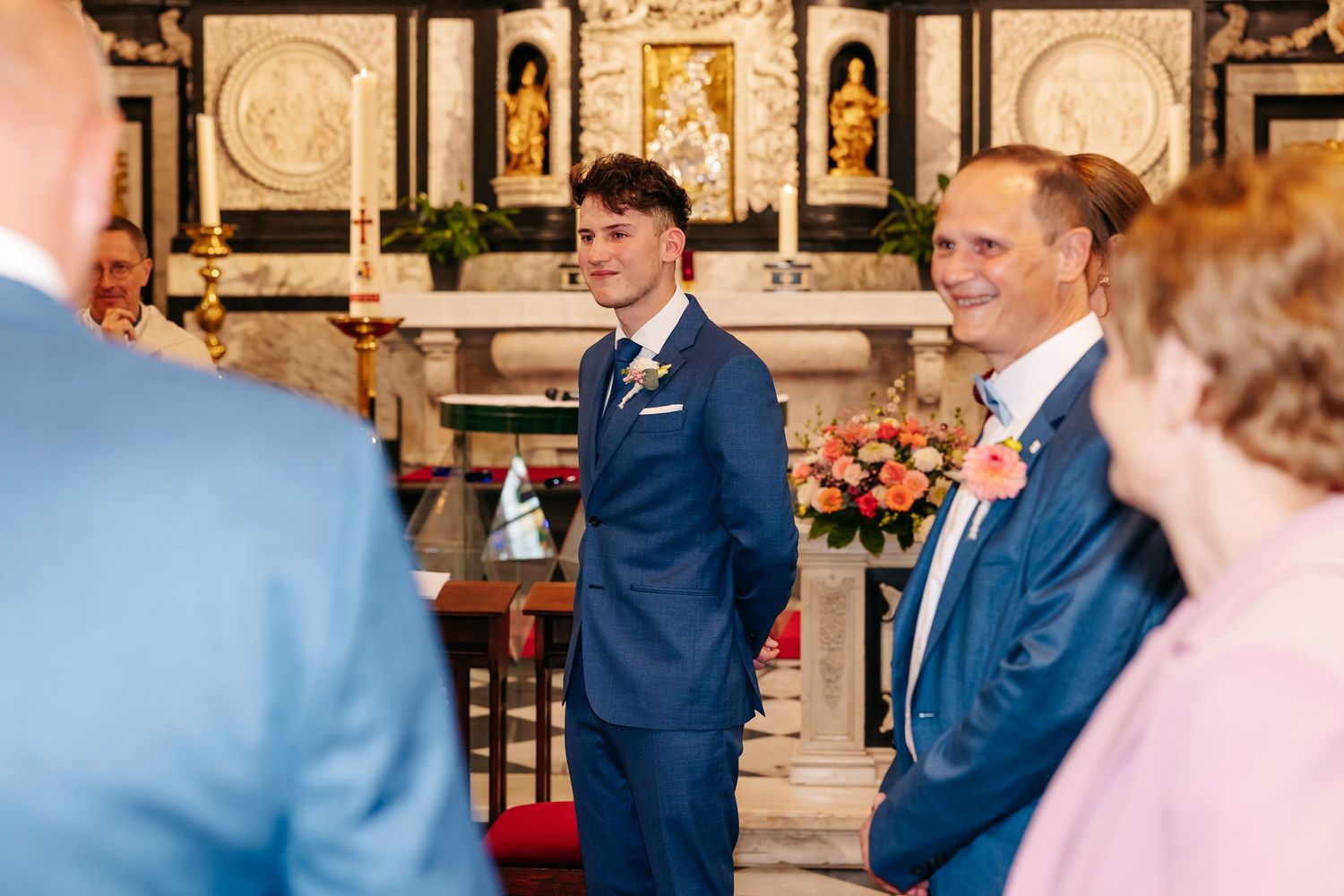 Groom in blue suit smiling at altar with guests in foreground inside an ornate decorated church.