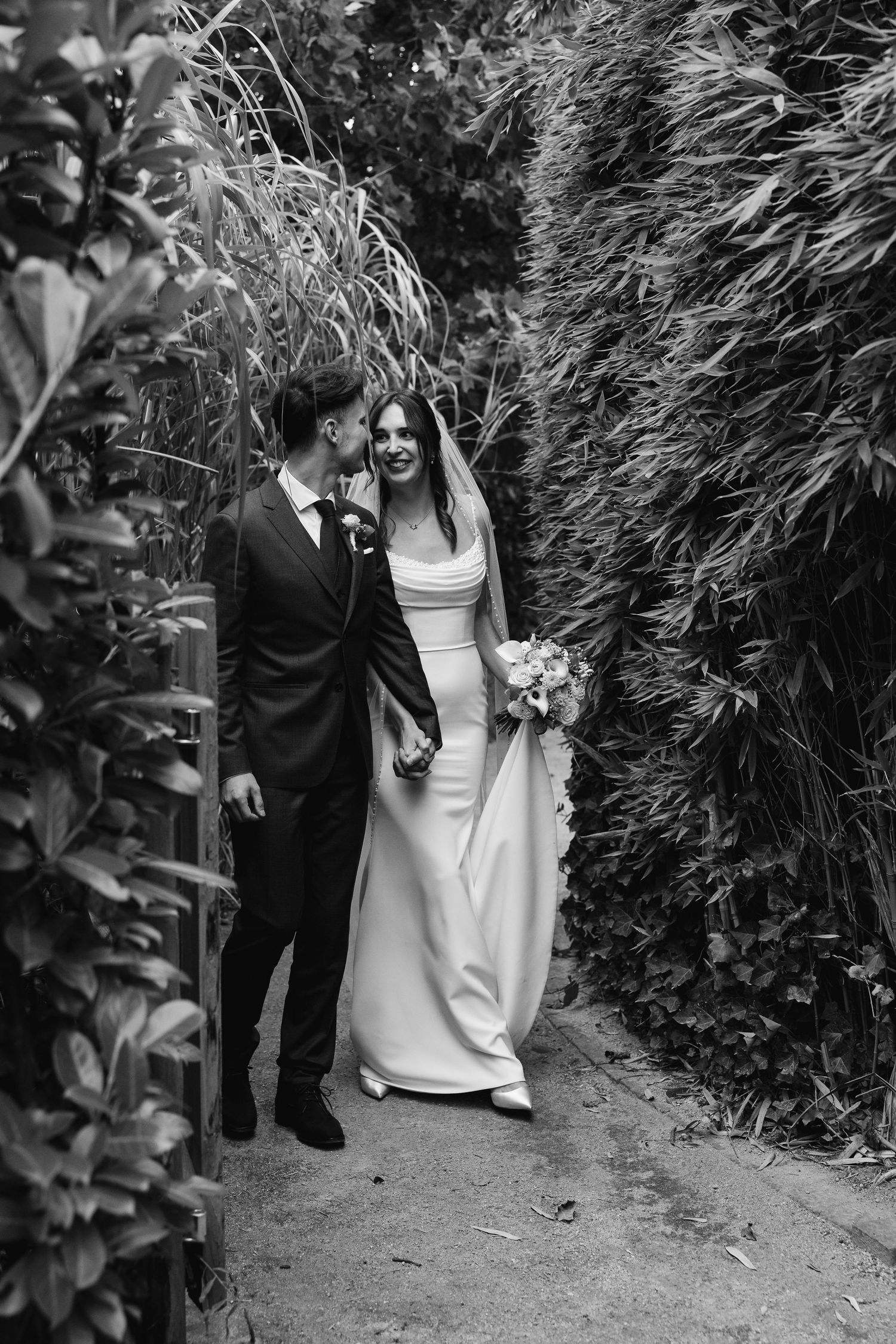 Black and white photo of bride and groom walking closely through a narrow garden pathway.