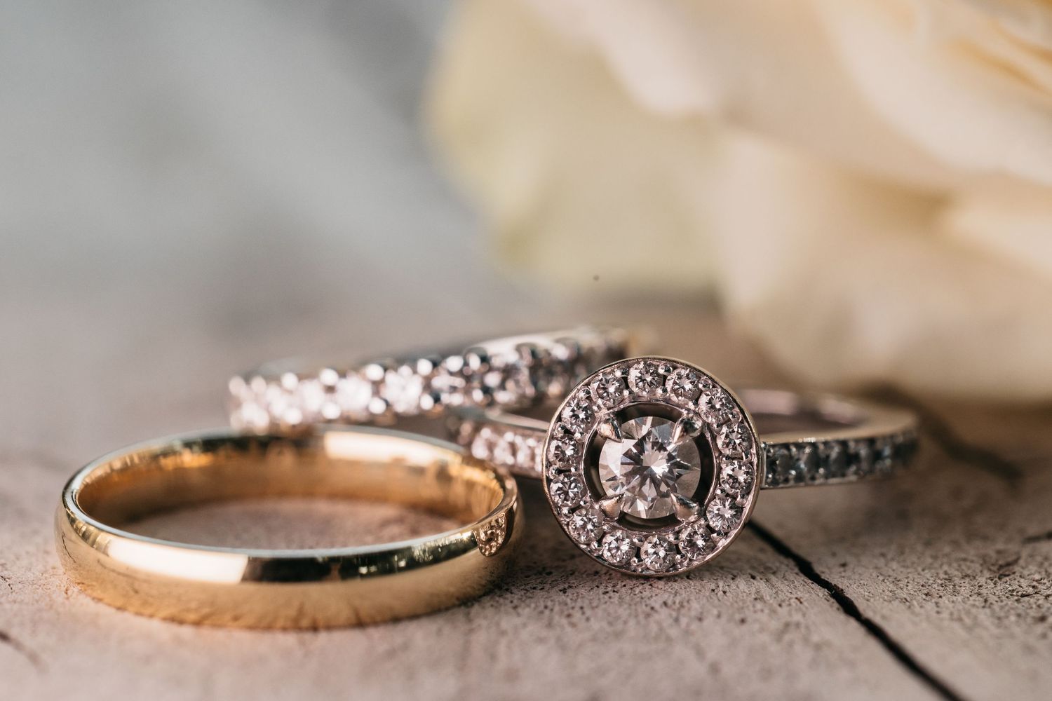 Close-up of diamond halo engagement ring and gold wedding band on rustic wooden surface.