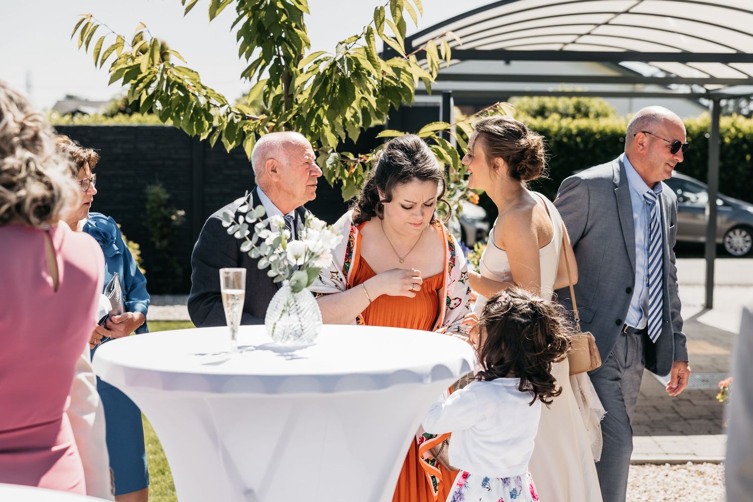 Wedding guests socializing outdoors at a white cocktail table in a sunny garden setting.