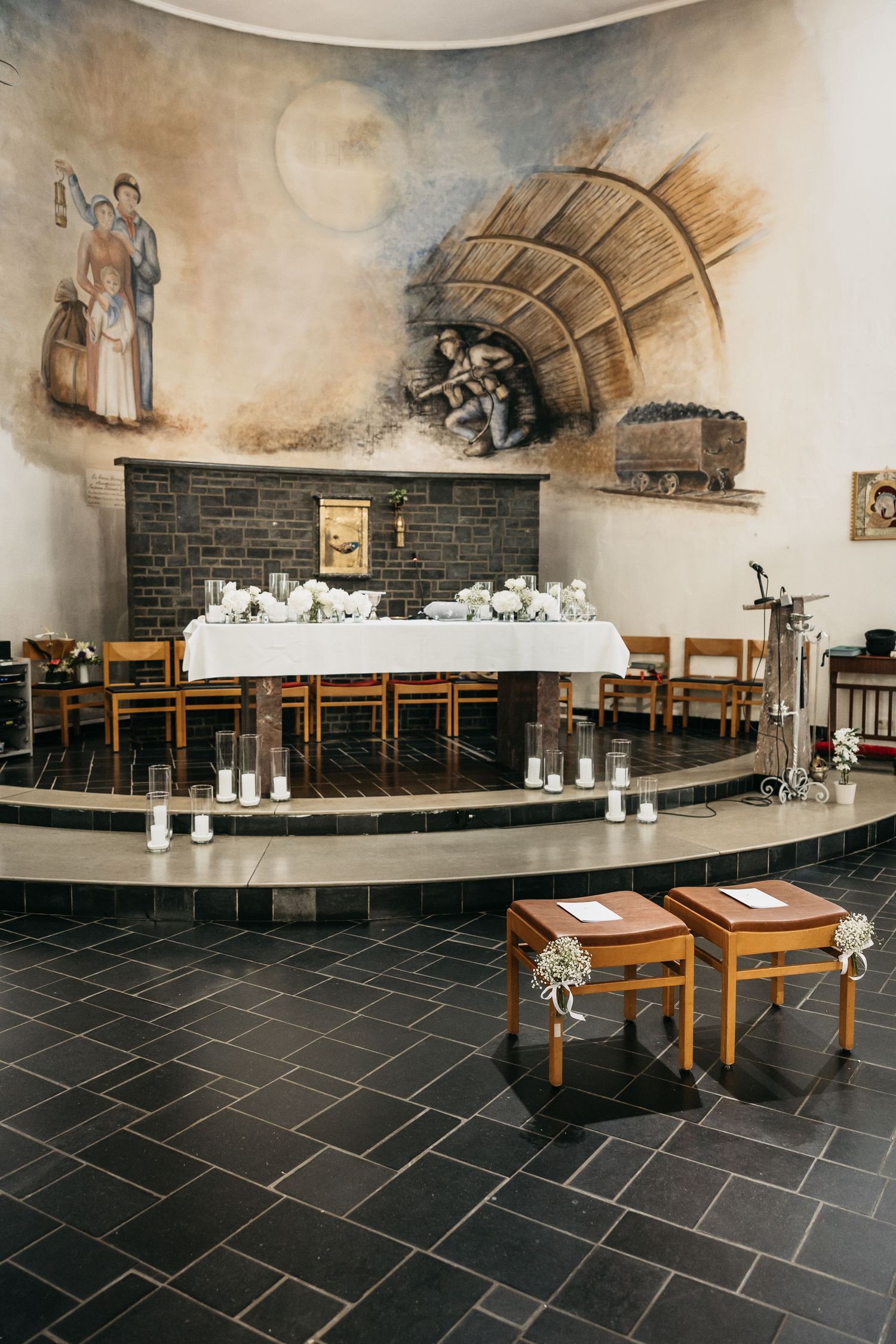Church altar with mining-themed fresco mural, white floral decorations, candles, and wooden kneeling stools.